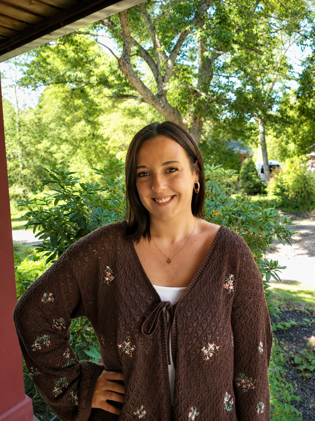 A smiling woman with shoulder-length dark hair stands outdoors in front of green leafy trees and plants, wearing a brown floral knit cardigan over a white top, in a sunny setting.