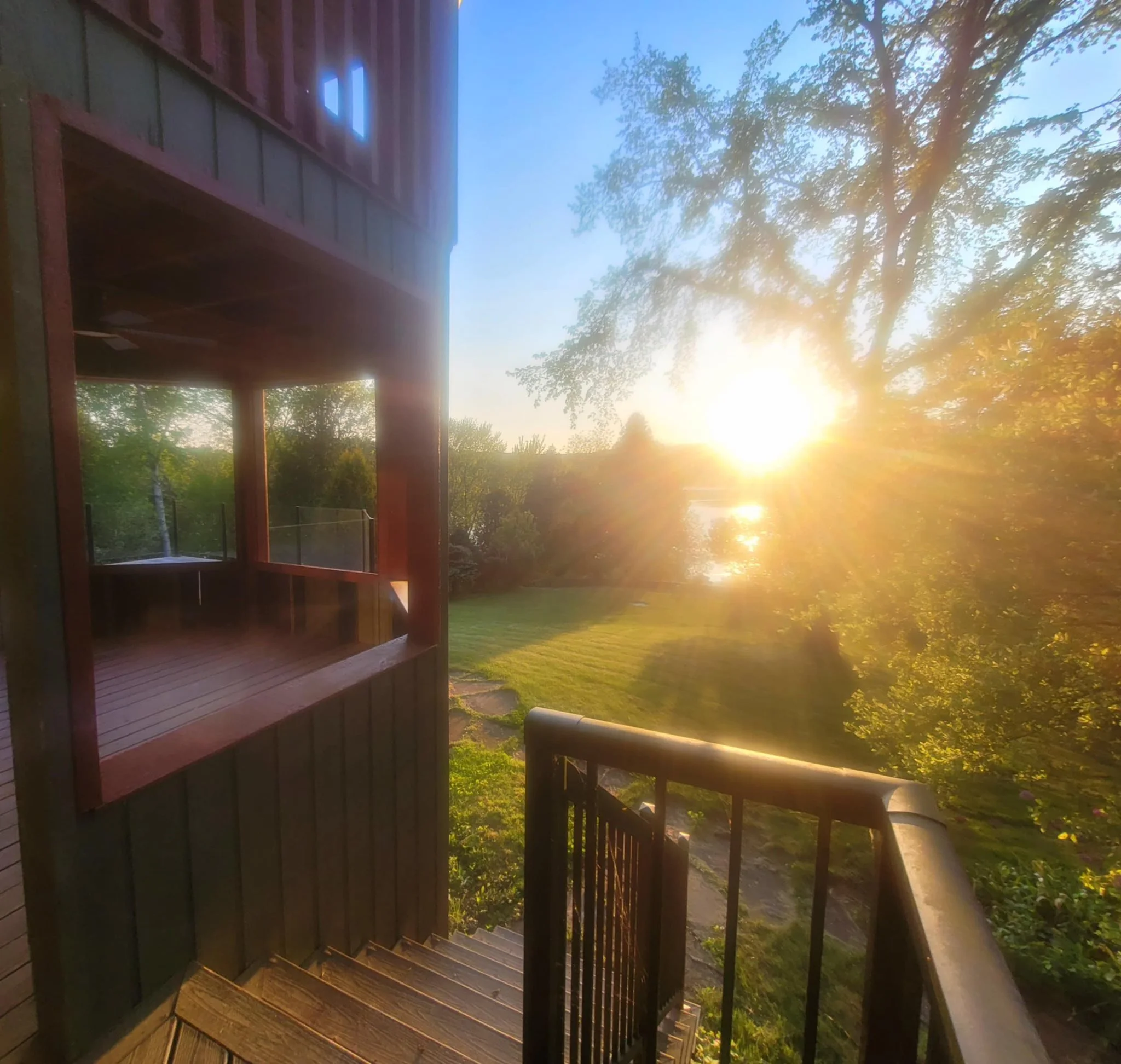 Sunset view from a staircase leading down to a grassy yard with trees and a pond, with sunlight filtering through the branches.