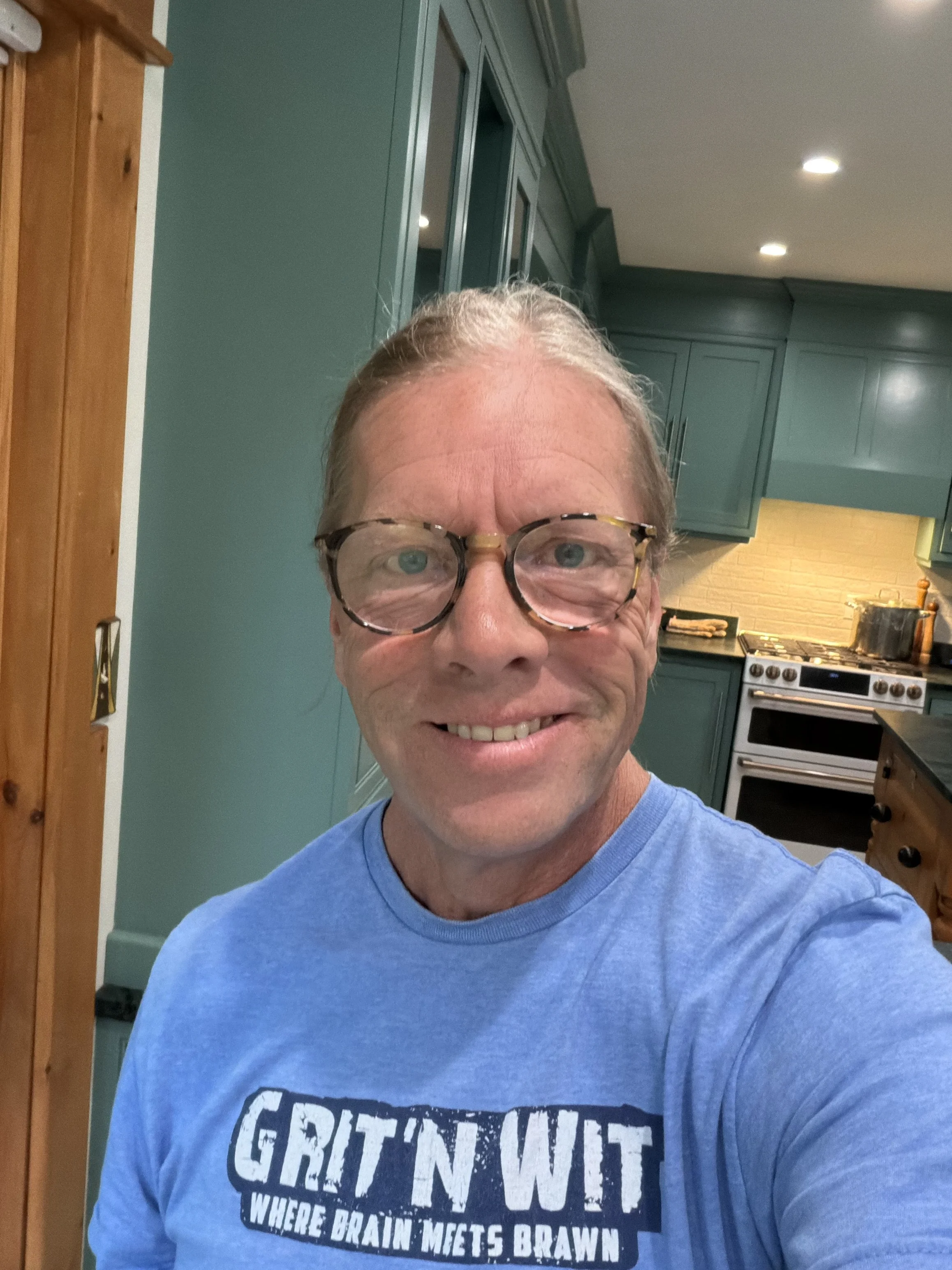 A man with glasses smiling in a kitchen with green cabinets and a yellow brick backsplash.