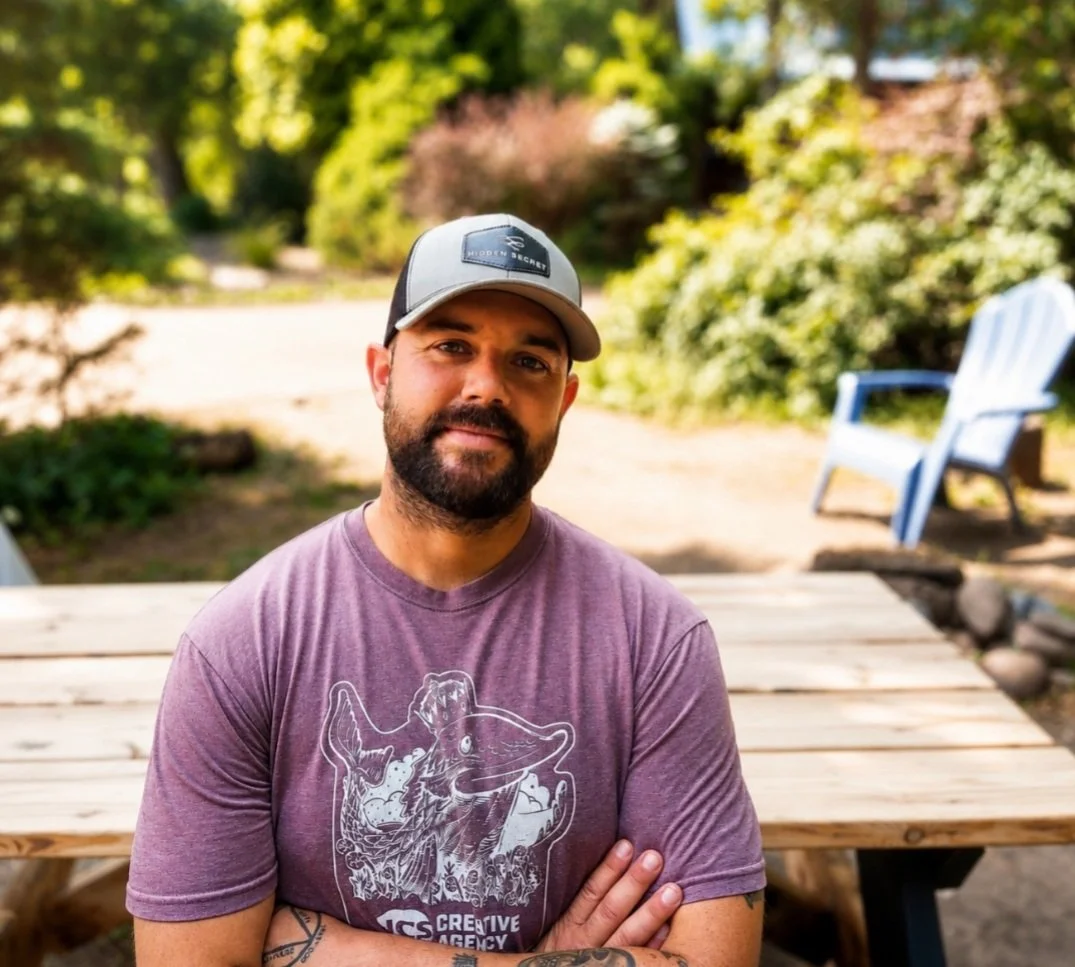 A man with a beard wearing a gray and black baseball cap and a purple T-shirt with a fish graphic, standing outdoors near a wooden table, with trees and a blue Adirondack chair in the background.