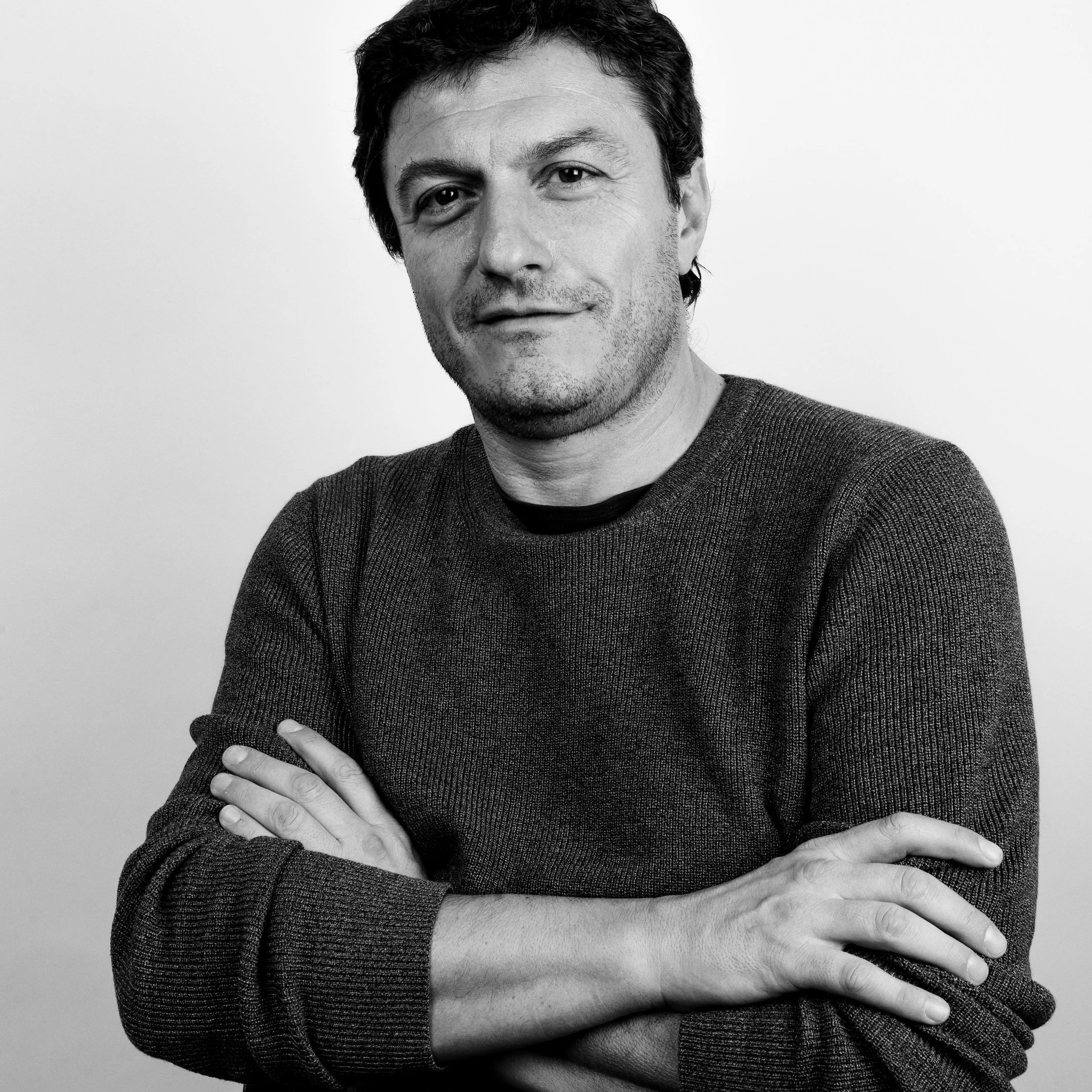 Black and white portrait of a man with dark hair, wearing a sweater, arms crossed, smiling slightly, against a plain background.