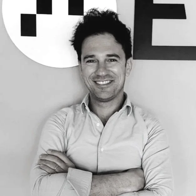 A smiling man with tousled hair and a beard, wearing a dark sweater, taking a selfie in front of a plain wall.