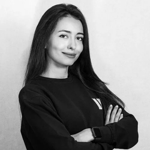 Black and white photo of a young woman with long dark hair, smiling slightly with arms crossed, wearing a dark sweatshirt and a smartwatch.