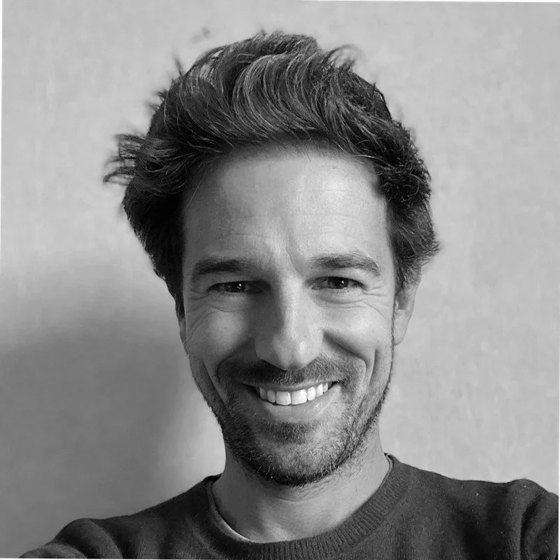 A smiling man with tousled hair and a beard, wearing a dark sweater, taking a selfie in front of a plain wall.
