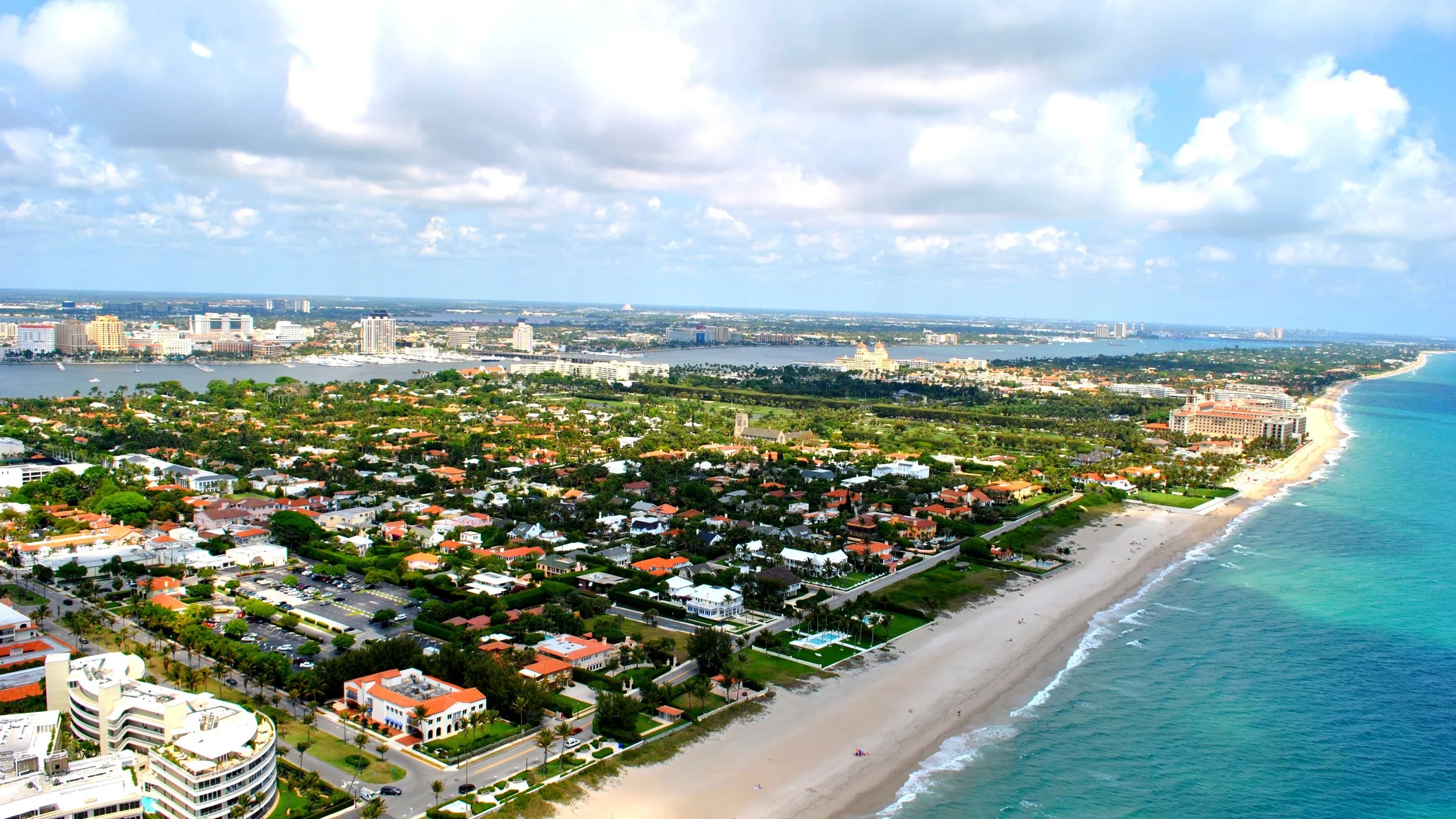 An aerial view of a tropical beach with white sand, turquoise water, and a coastal town with houses and taller buildings in the background under a partly cloudy sky.
