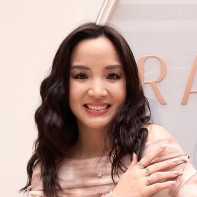 A woman with long, wavy dark hair smiling at the camera, wearing a light-colored top and rings on her fingers.
