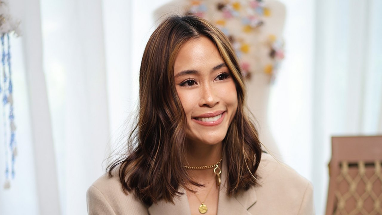 A woman with shoulder-length brown wavy hair, smiling, wearing a beige blazer and gold jewelry, in an indoor setting with light-colored curtains and a floral lamp in the background.