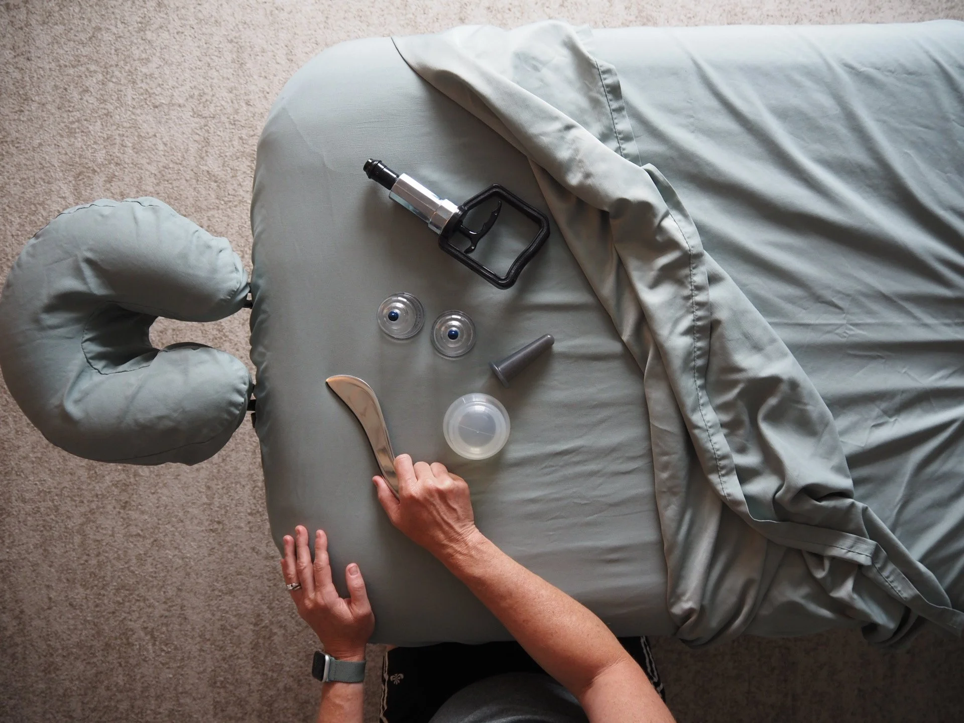 A person lying face down on a massage table with a gray sheet. Another person is preparing to administer a massage or medical treatment, holding a metal spoon-like tool. Various medical or massage tools are spread out on the table, including a spray bottle, small containers, and suction cups.
