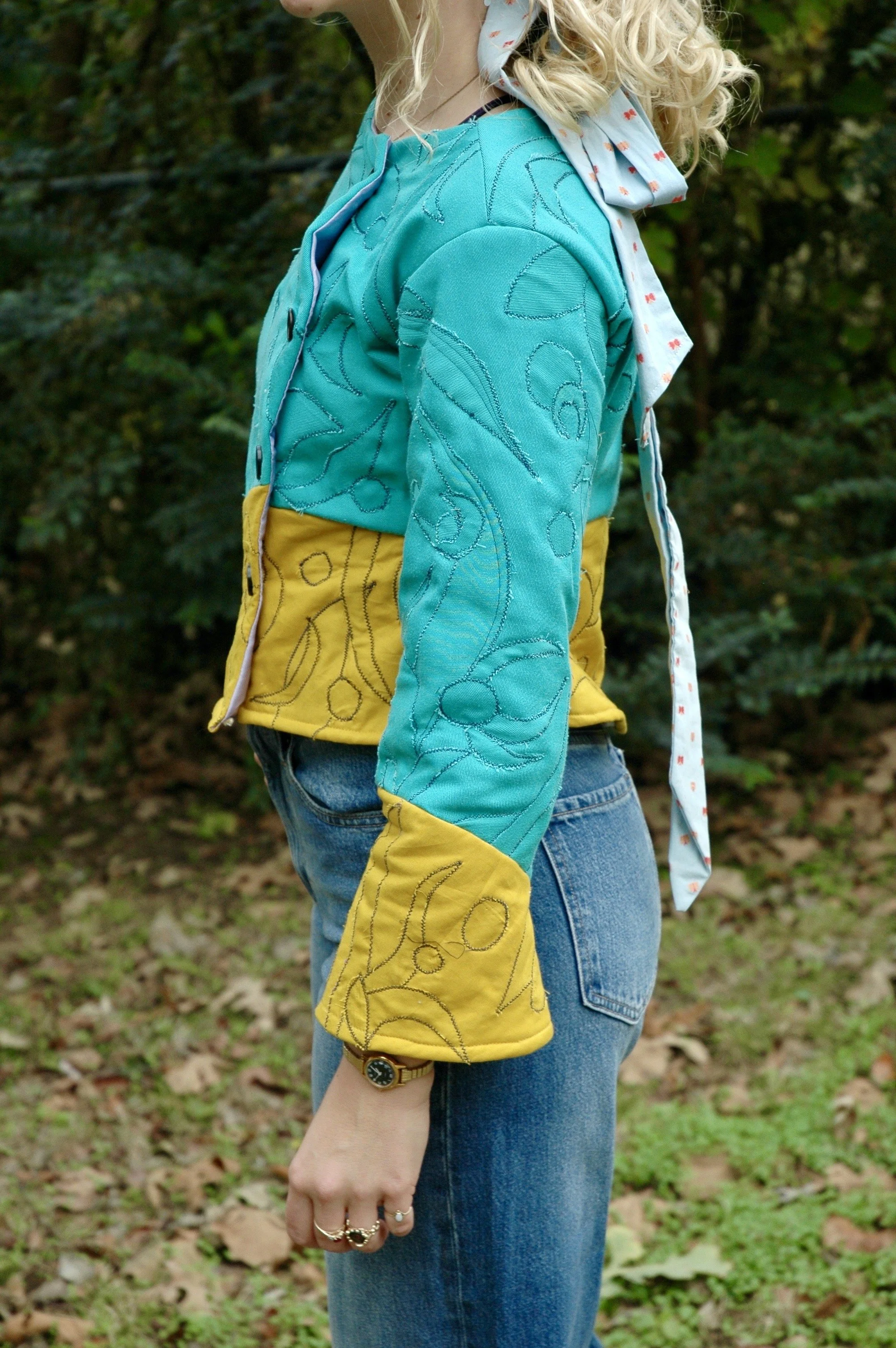 A person wearing a turquoise quilted jacket with yellow patches on the sleeves, blue jeans, and a white scarf with red patterns, standing outdoors with green foliage and fallen leaves in the background. Part of their face and head are not visible.