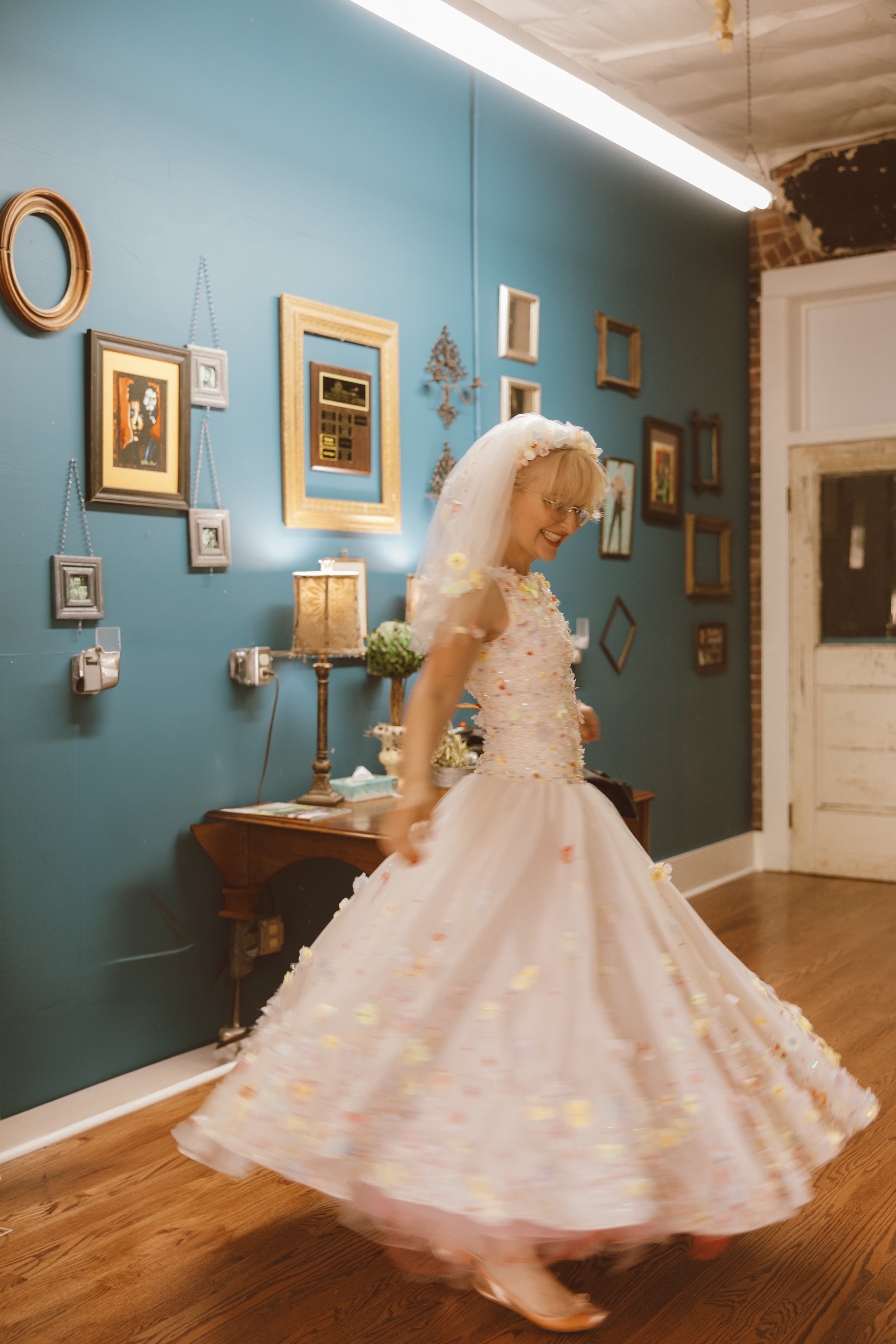 A woman dressed in a floral, pastel-colored gown with a matching veil is spinning in a dance or celebration, creating a flowing motion of her dress in a room with blue walls, decorated with picture frames, lamps, and artwork.