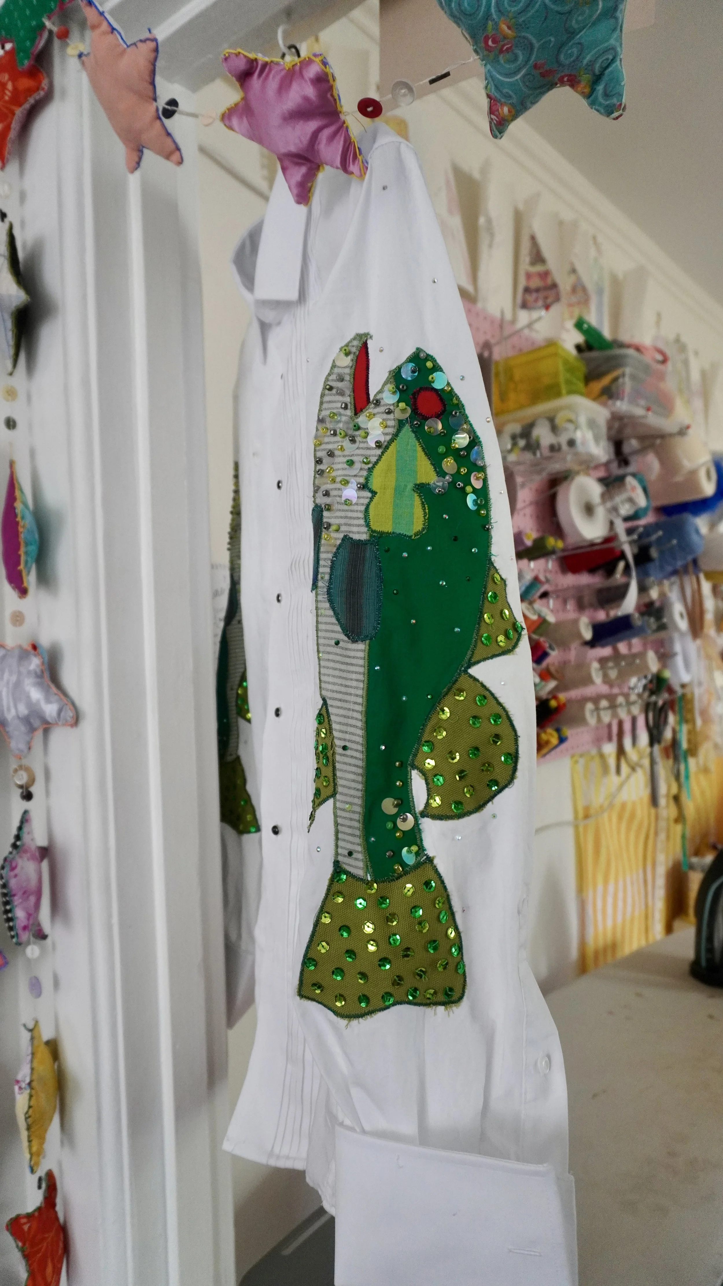A fabric applique of a green and yellow fish with sequins and rhinestones, sewn onto a white shirt, hanging on a line with star-shaped plush toys and a background of craft supplies and storage bins.