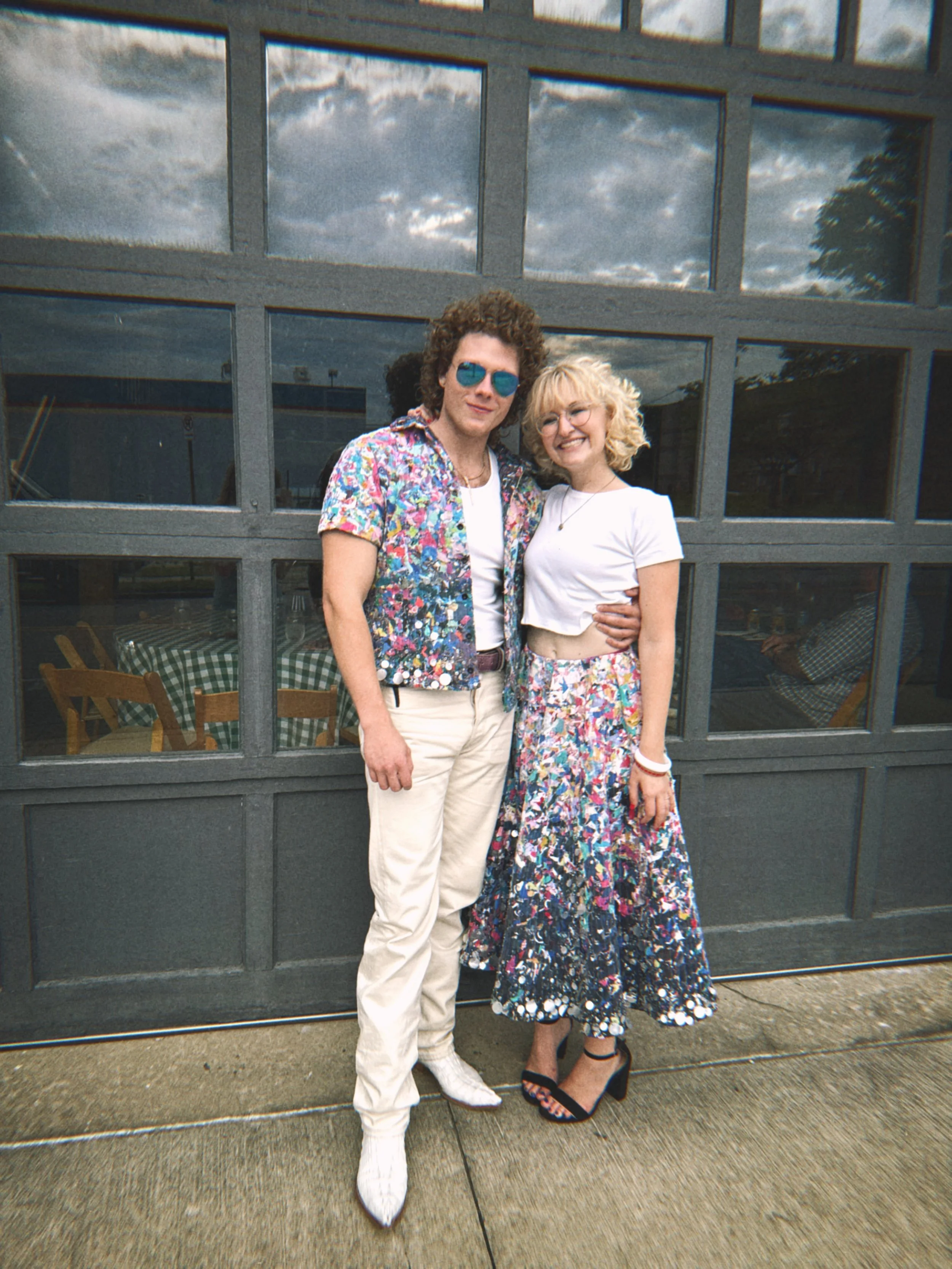 A man and woman standing together outdoors, smiling, in front of a modern building with large windows reflecting a cloudy sky. The woman is wearing a white t-shirt and a colorful floral skirt, black high heels, and glasses. The man is wearing a color