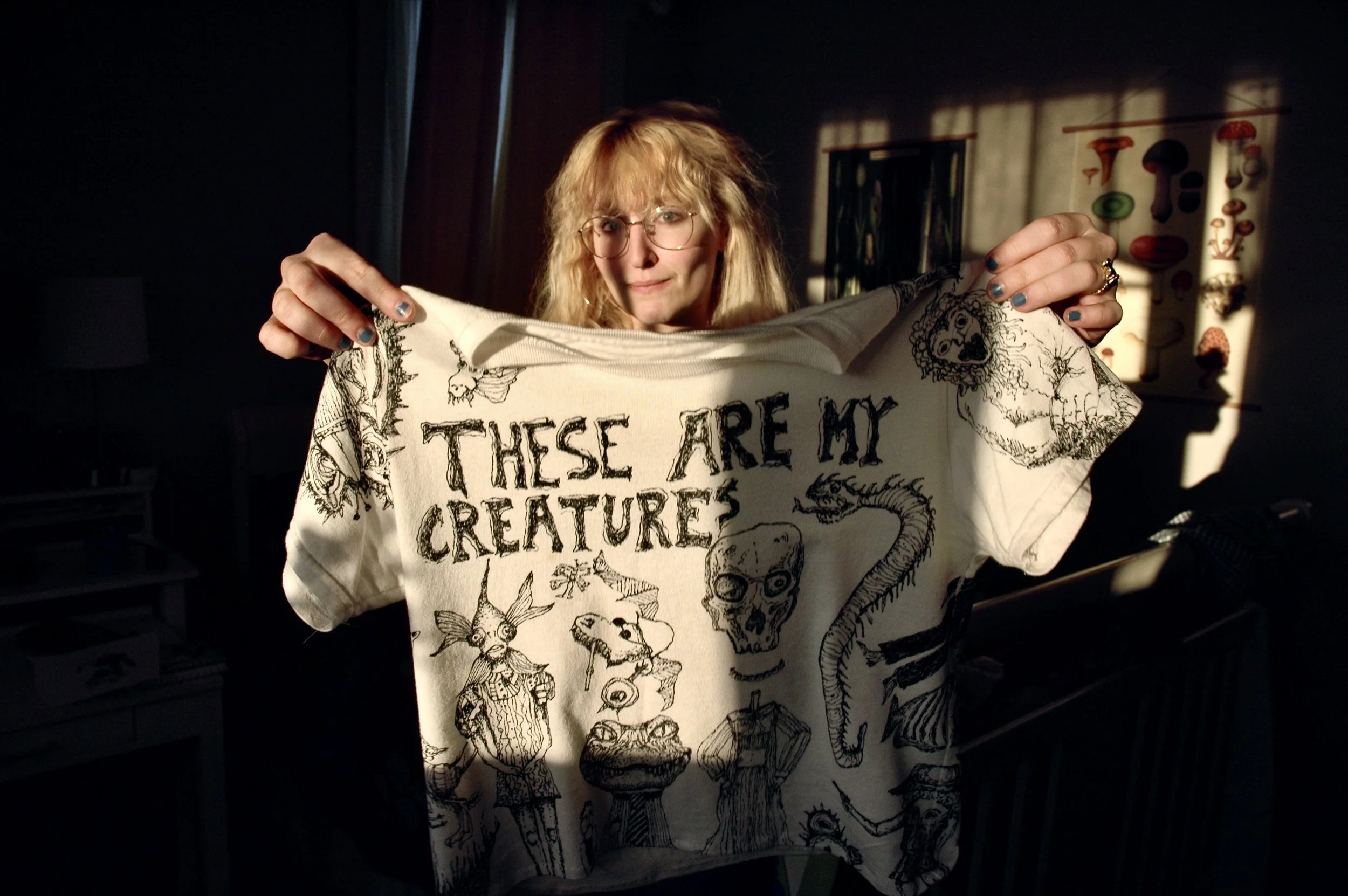 A person with blonde hair and glasses holding up a white shirt with black illustrations of animals and skulls, and the phrase 'These are my creatures' written on it.