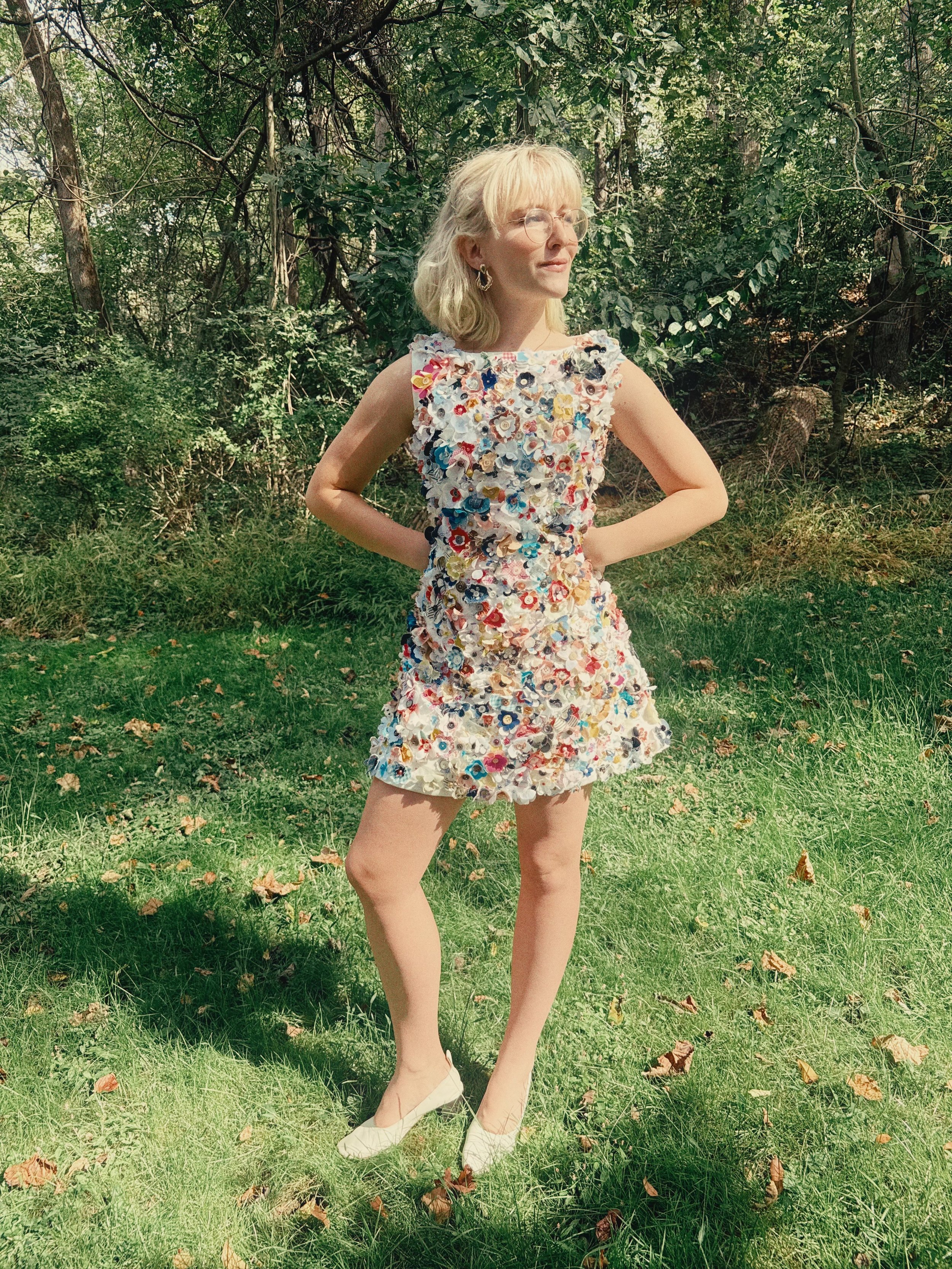 Woman wearing a dress made of various colorful fabric flowers standing on grass in a lush green outdoor setting.
