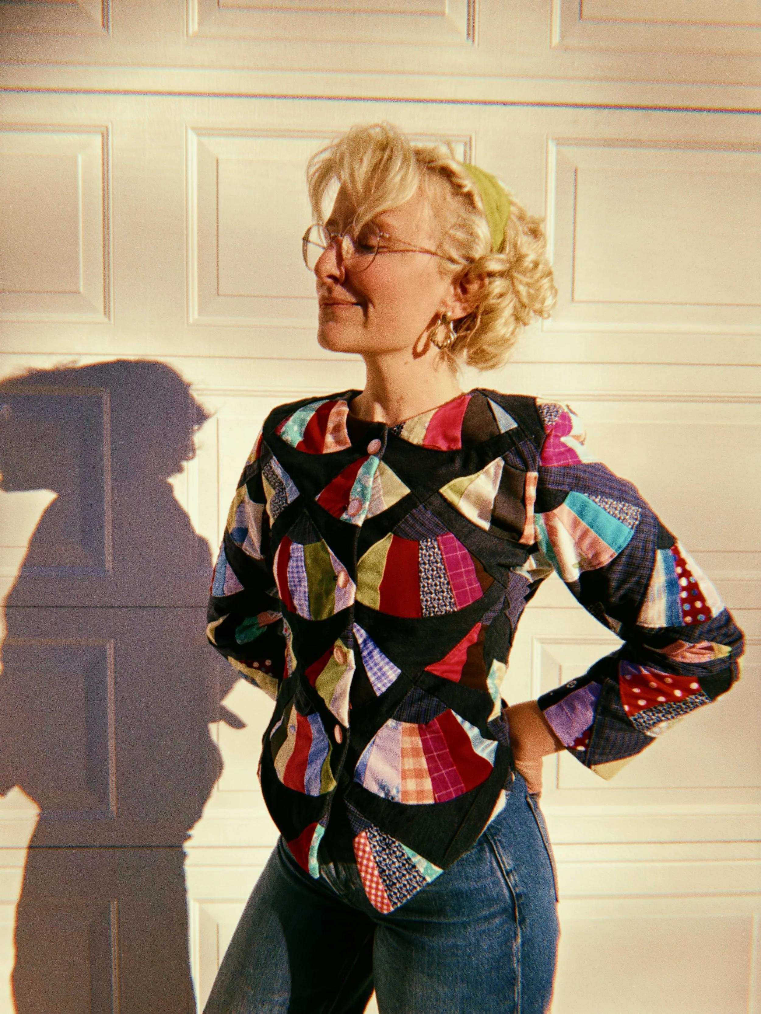 A woman with blonde curly hair tied with a green band, wearing glasses and hoop earrings, stands sideways in front of a beige paneled wall, dressed in a colorful patchwork shirt and dark jeans.