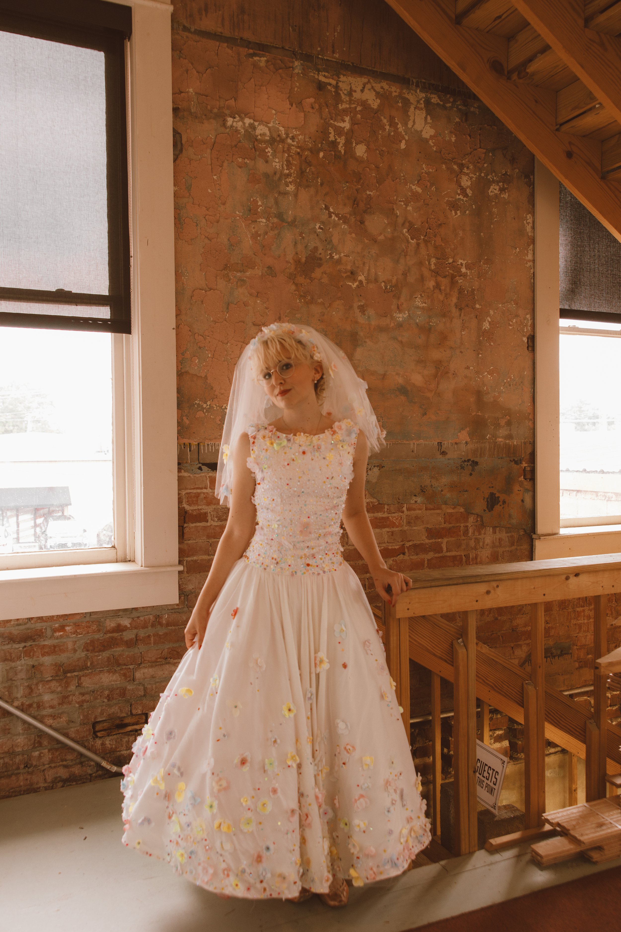A woman dressed in a white gown with colorful floral details, wearing a veil and glasses, standing indoors by a brick wall and windows.