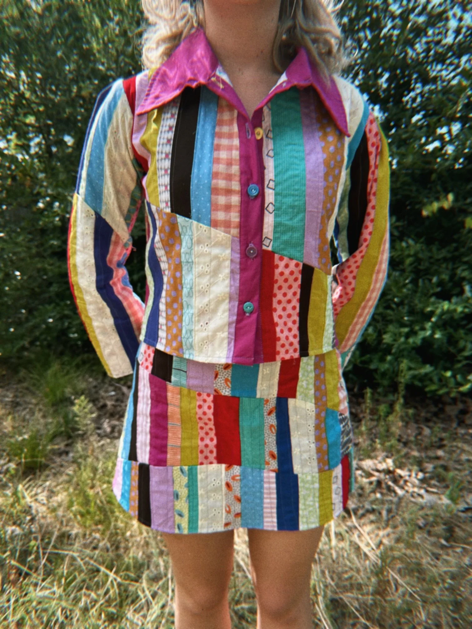 A person wearing a colorful patchwork dress with various patterns and colors, standing outdoors among green foliage and grass.