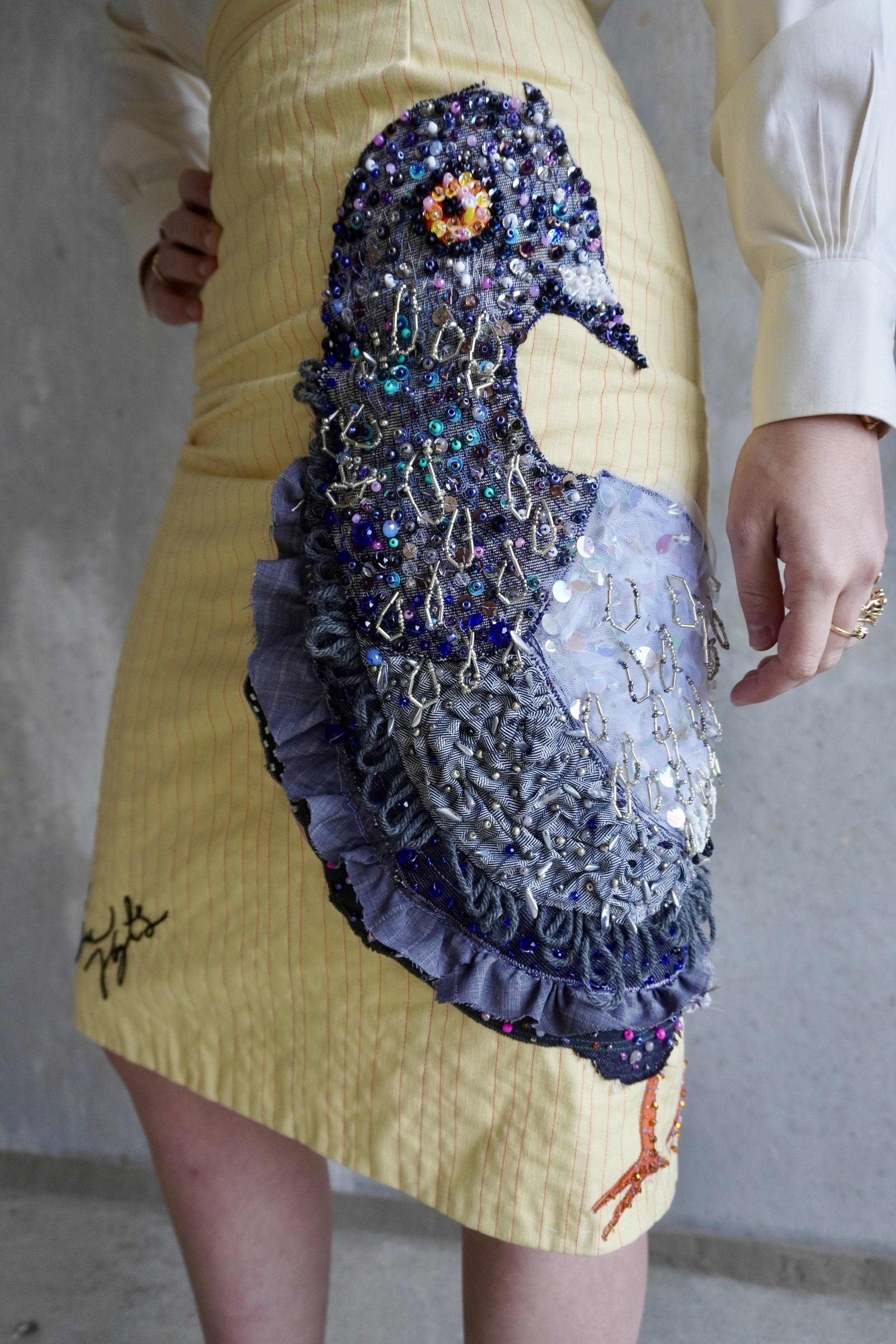 A person wearing a yellow skirt with an intricate beaded patchwork design featuring various beads, jewels, and fabric embellishments, including a partial orange embroidered design.