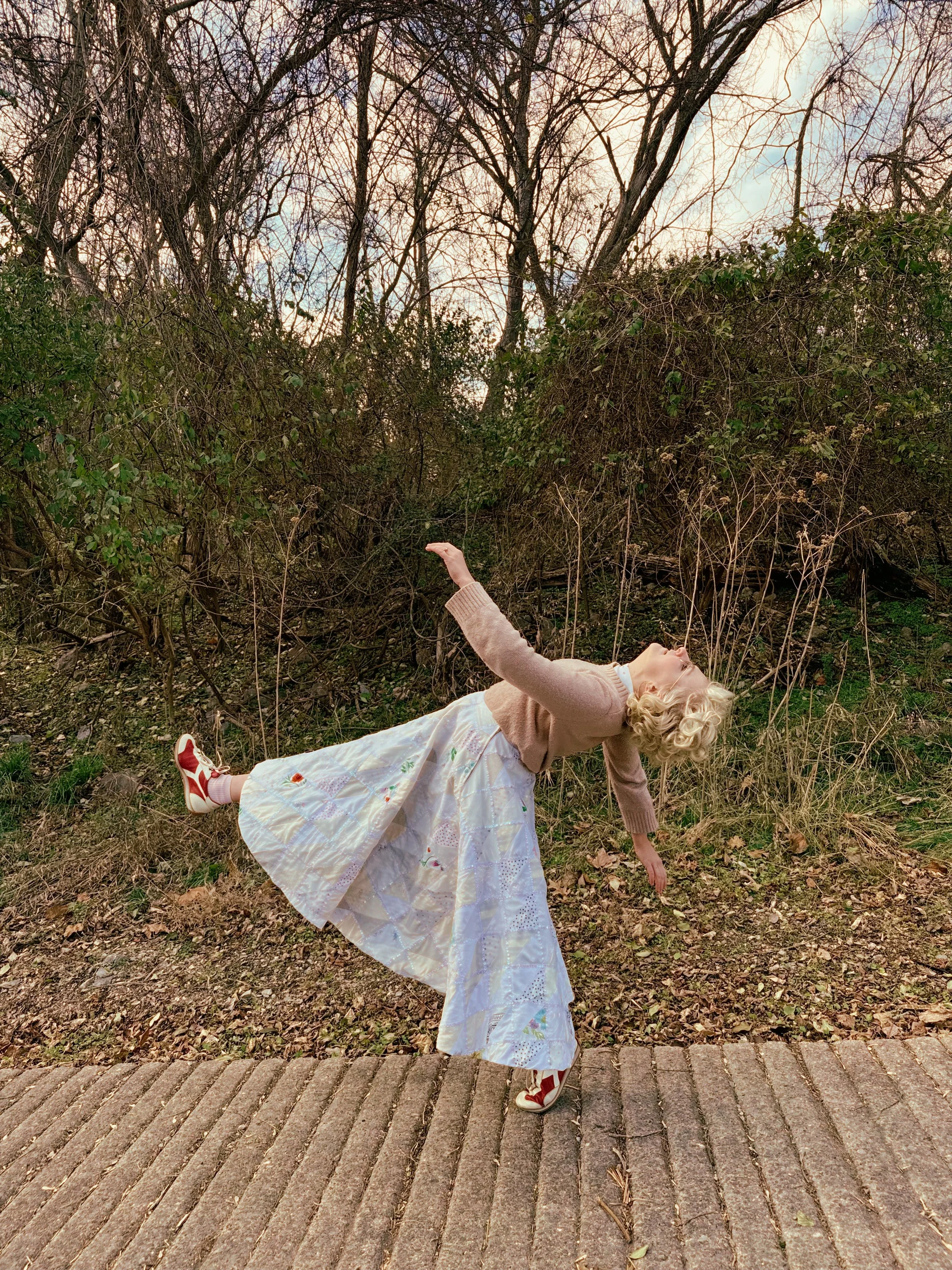 A girl with blonde, curly hair laying on her back in a wooded area, with her left arm extended upward and her right arm down toward the ground, wearing a dress with a geometric pattern, a beige cardigan, and red and white sneakers, on a sidewalk with