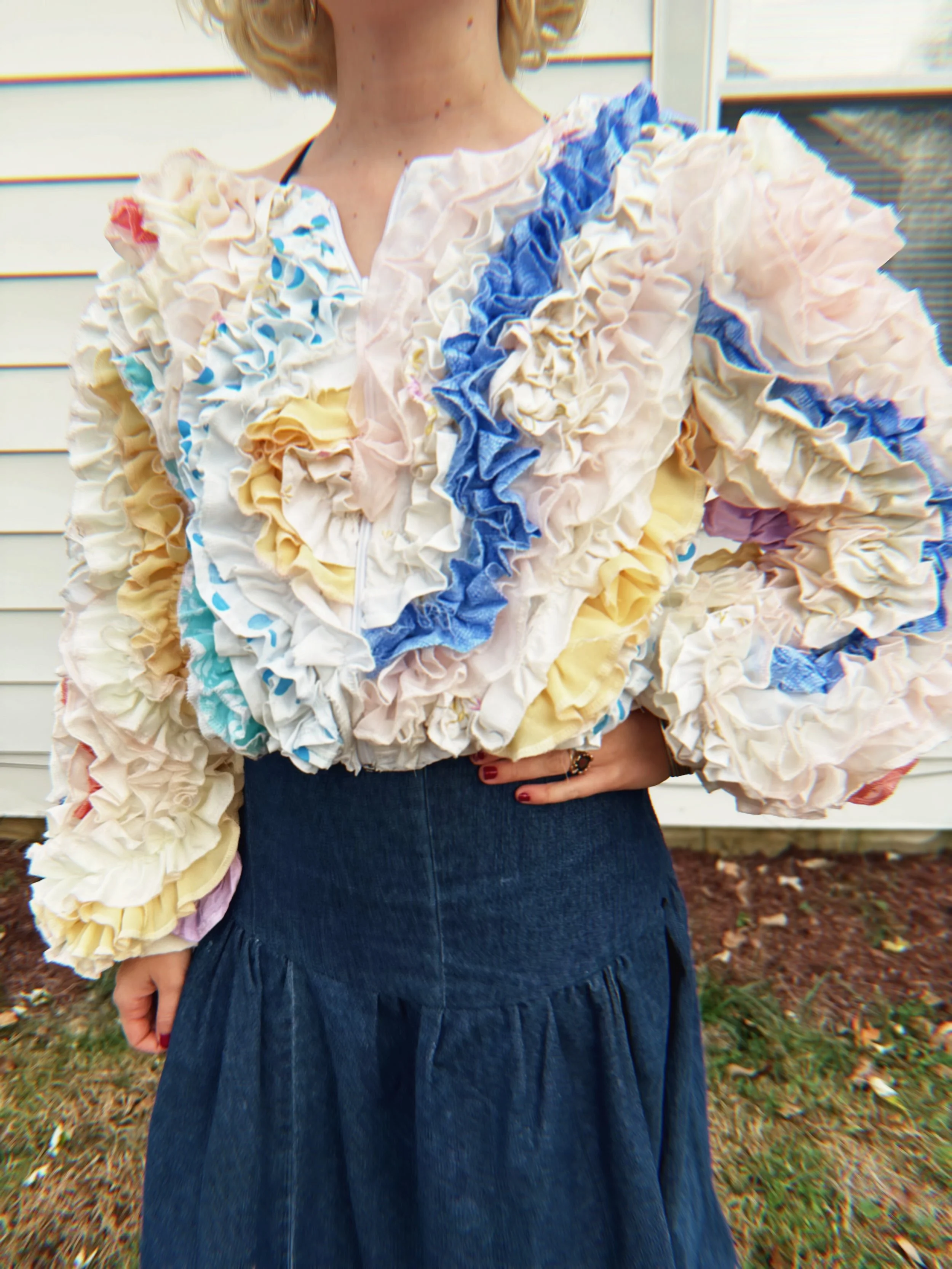 Person wearing a frilly multicolored sweater and a dark blue denim skirt, standing outside in front of a house with horizontal siding, holding their hand on their waist.