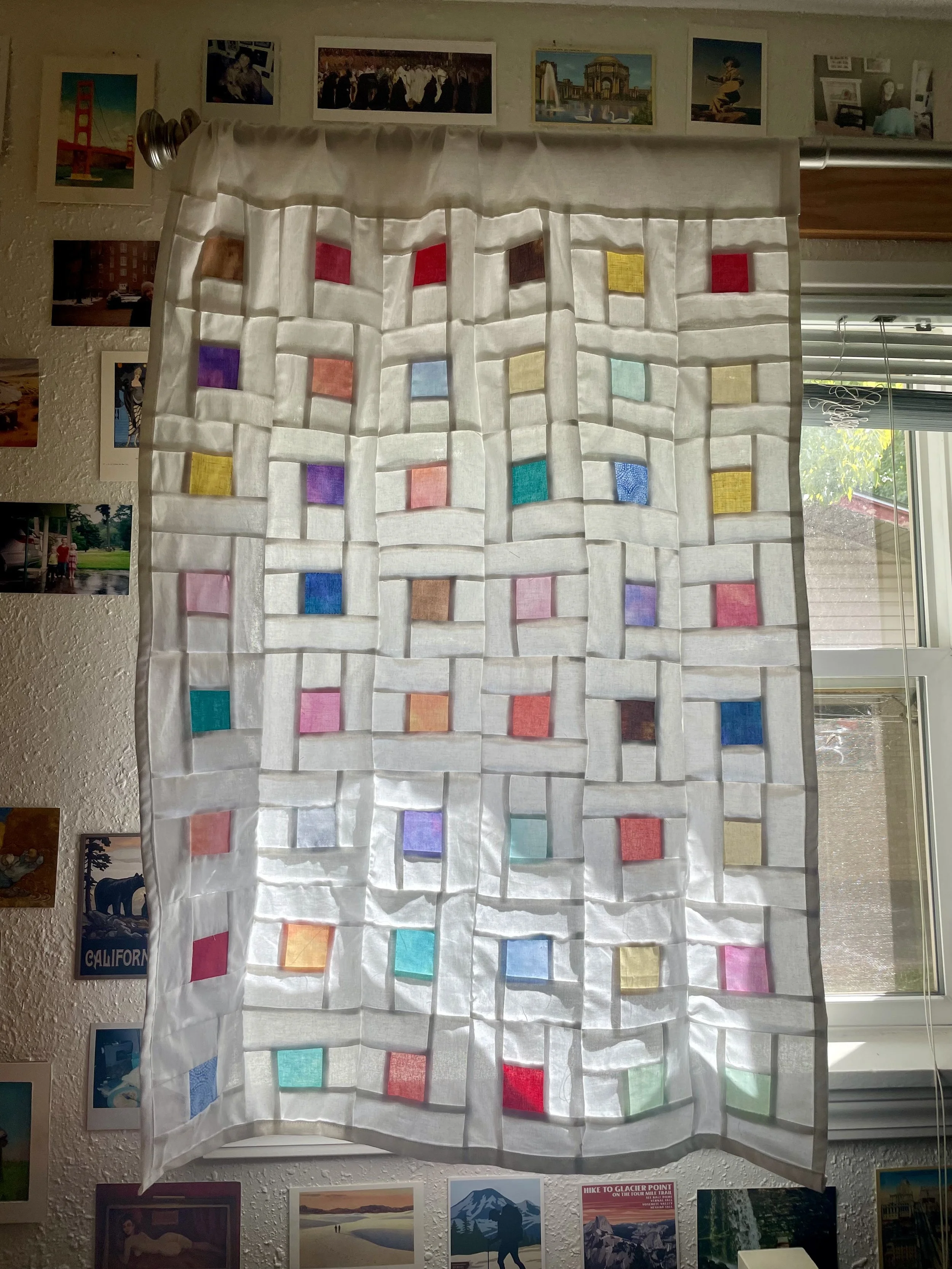 Colorful fabric squares arranged in a grid pattern behind a sheer white curtain, with small images and photographs on the wall around it and a window with blinds to the right.
