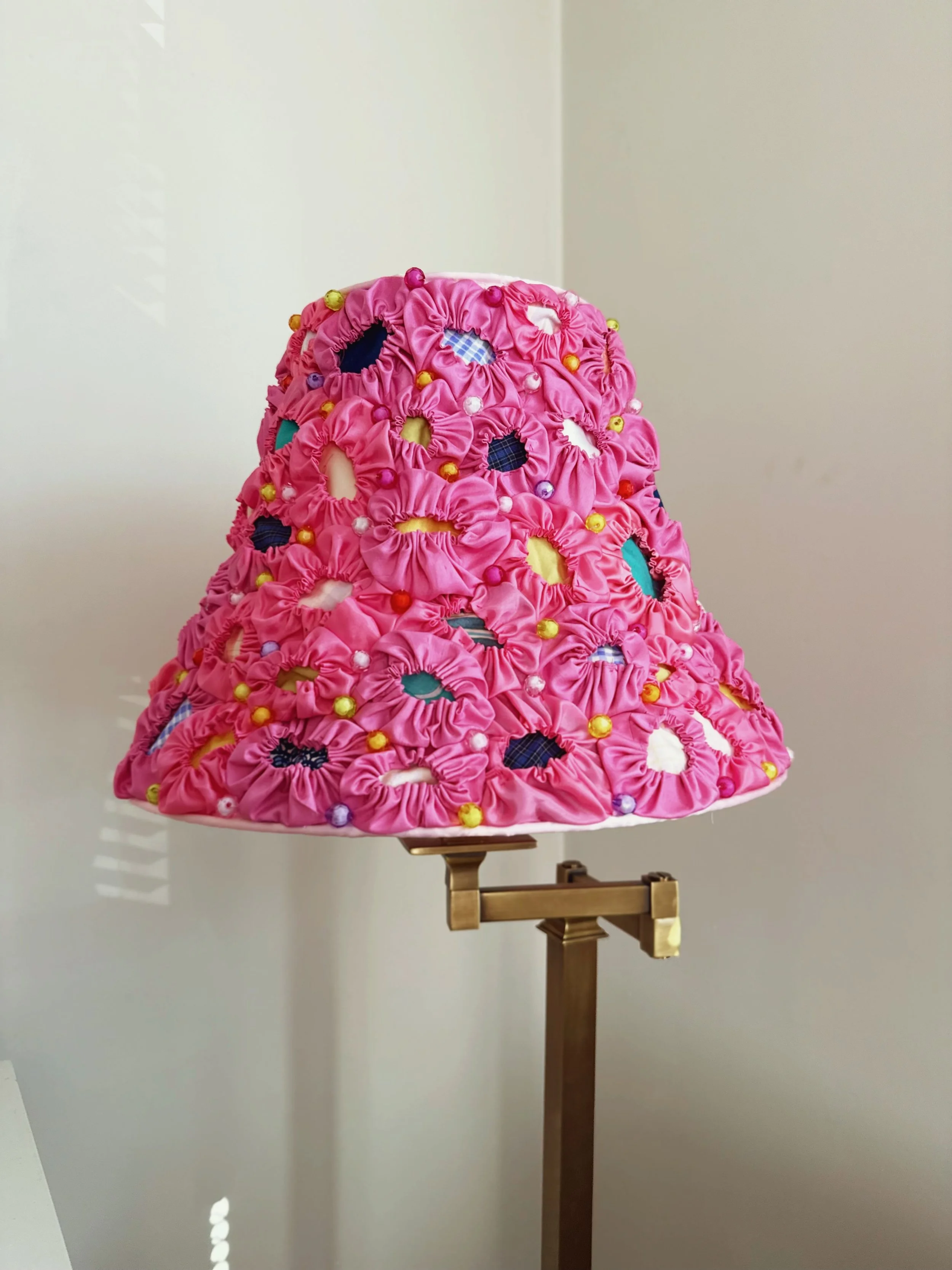 A decorative lampshade made of pink fabric with gathered ruffles, decorated with colorful small beads and patches with floral and checkered patterns.