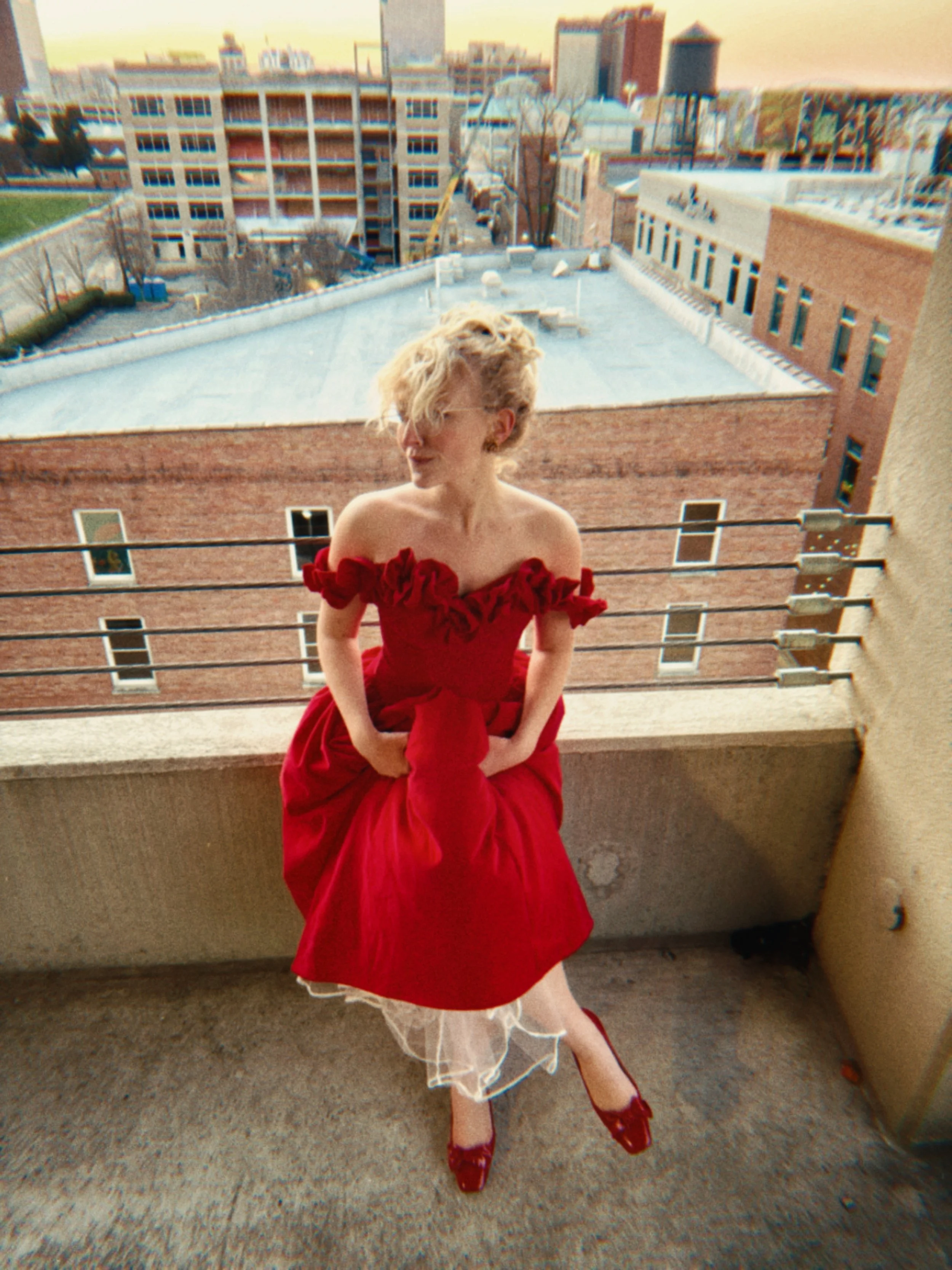 A woman in a red off-the-shoulder dress with ruffle details and a tulle underskirt standing on a balcony. She is wearing red shoes and sunglasses, with her curly blonde hair partially covering her face. The city skyline and rooftops are visible in th