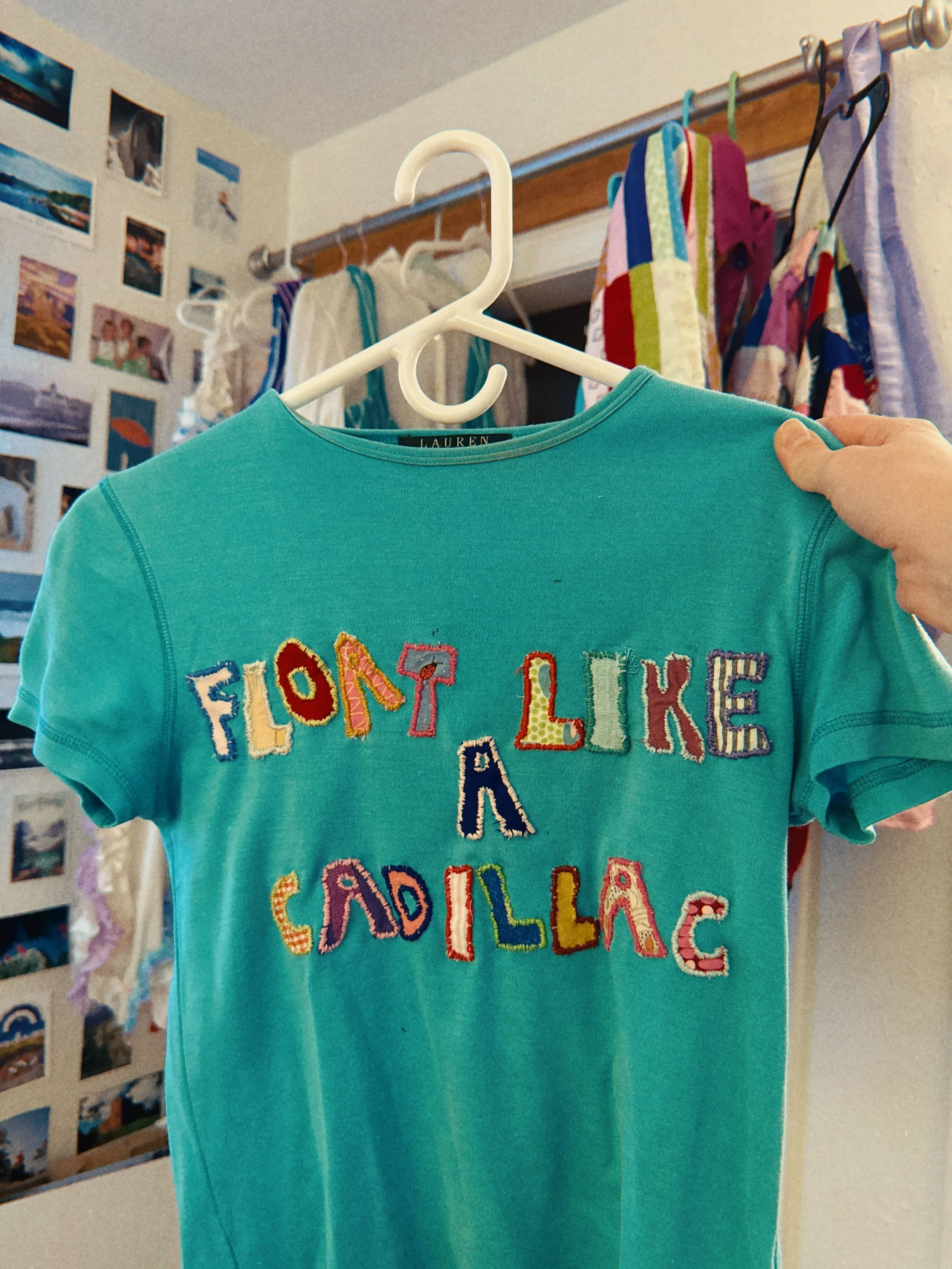 A turquoise t-shirt with colorful embroidered words, held on a white hanger, in front of a wall with photo collages and hanging clothes.