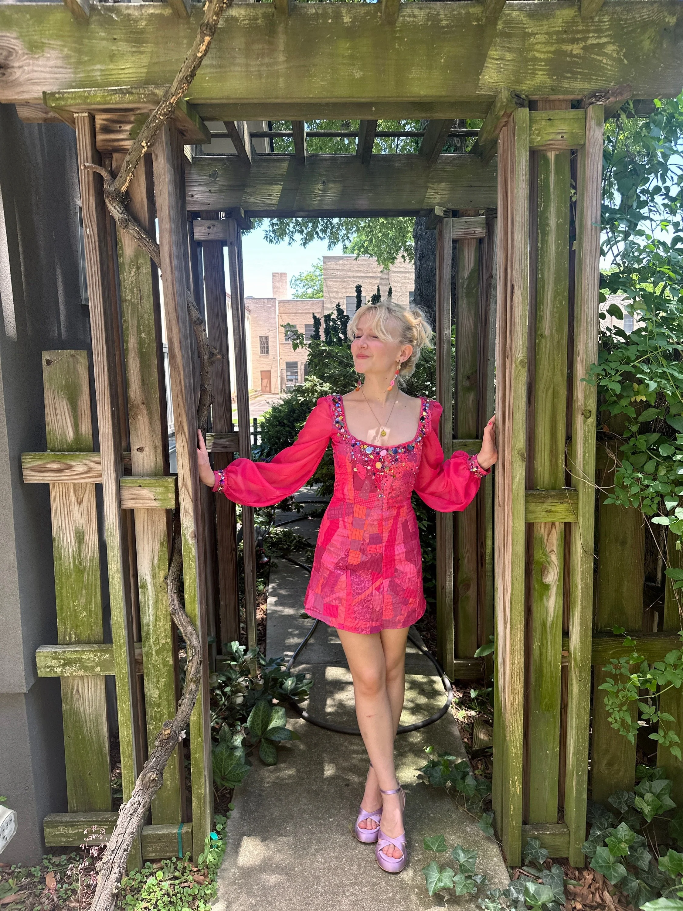 A woman with light blonde hair standing in a wooden archway outdoors, wearing a bright pink dress and pale pink platform shoes, with her eyes closed and her right hand touching the wood.