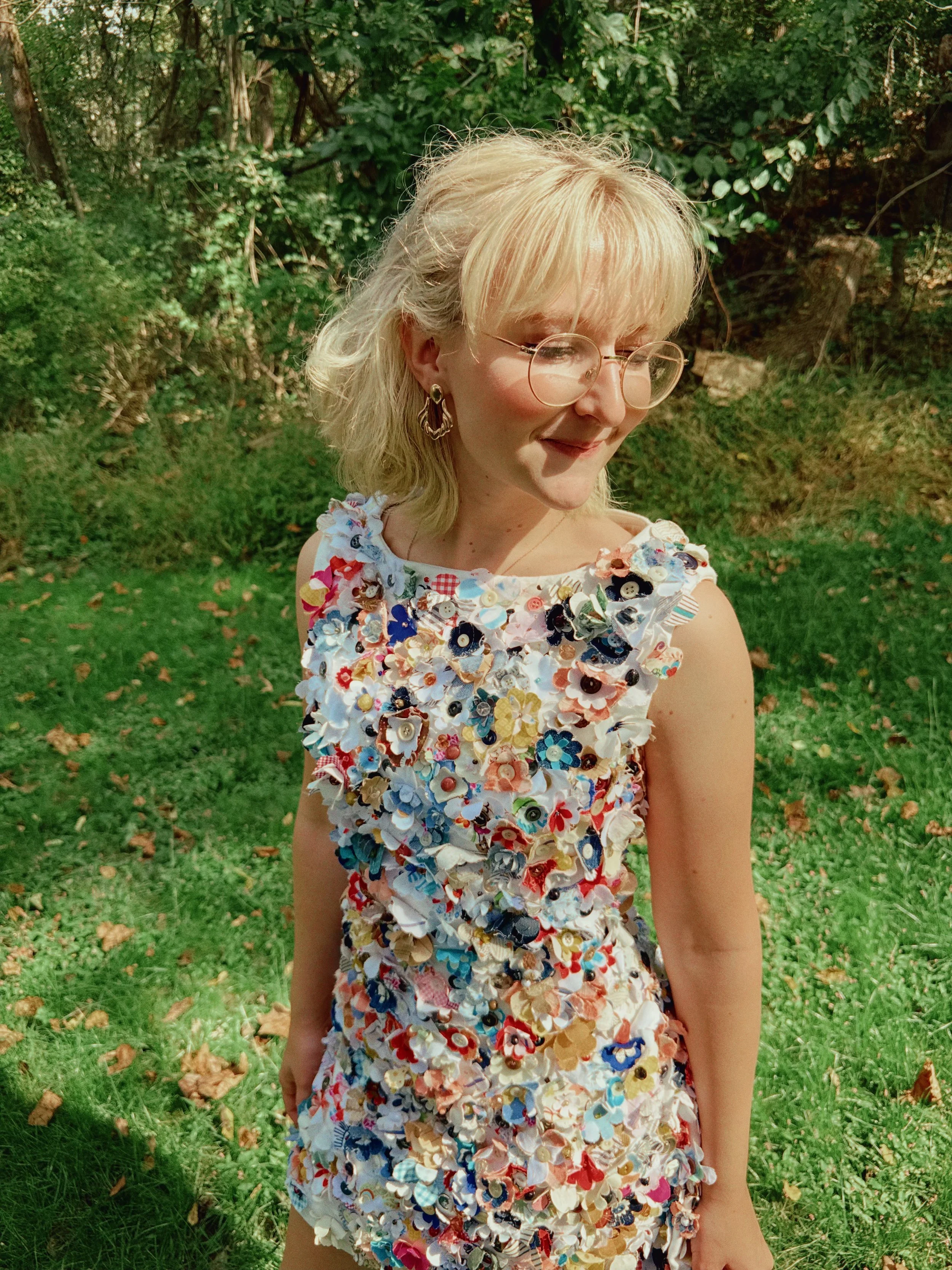 A woman with blonde hair and glasses smiling outdoors in a green park, wearing a dress decorated with colorful buttons and fabric flowers.