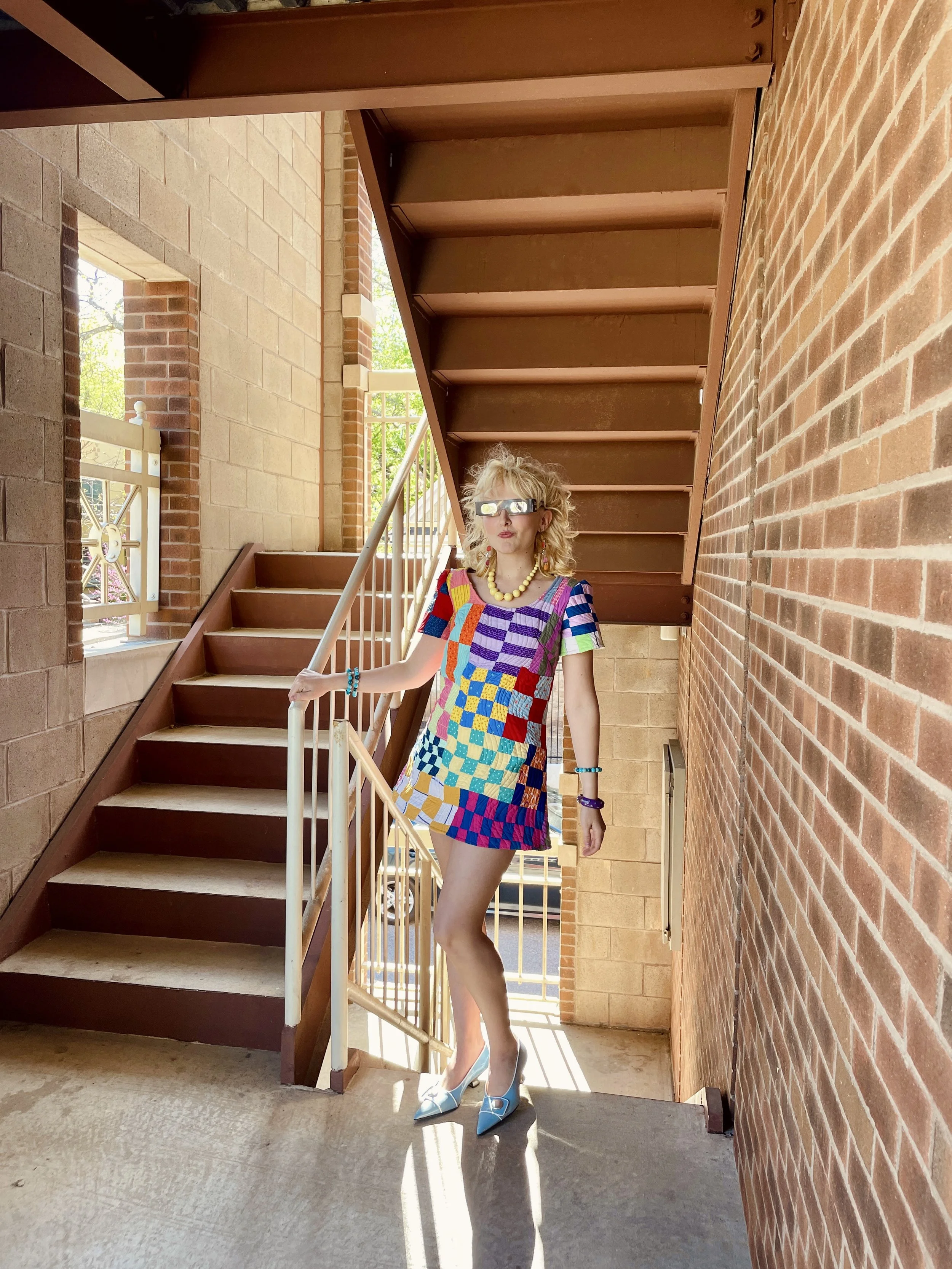 Woman wearing a colorful patchwork dress, high heels, and large reflective sunglasses standing on an outdoor staircase with brick walls and a railing.