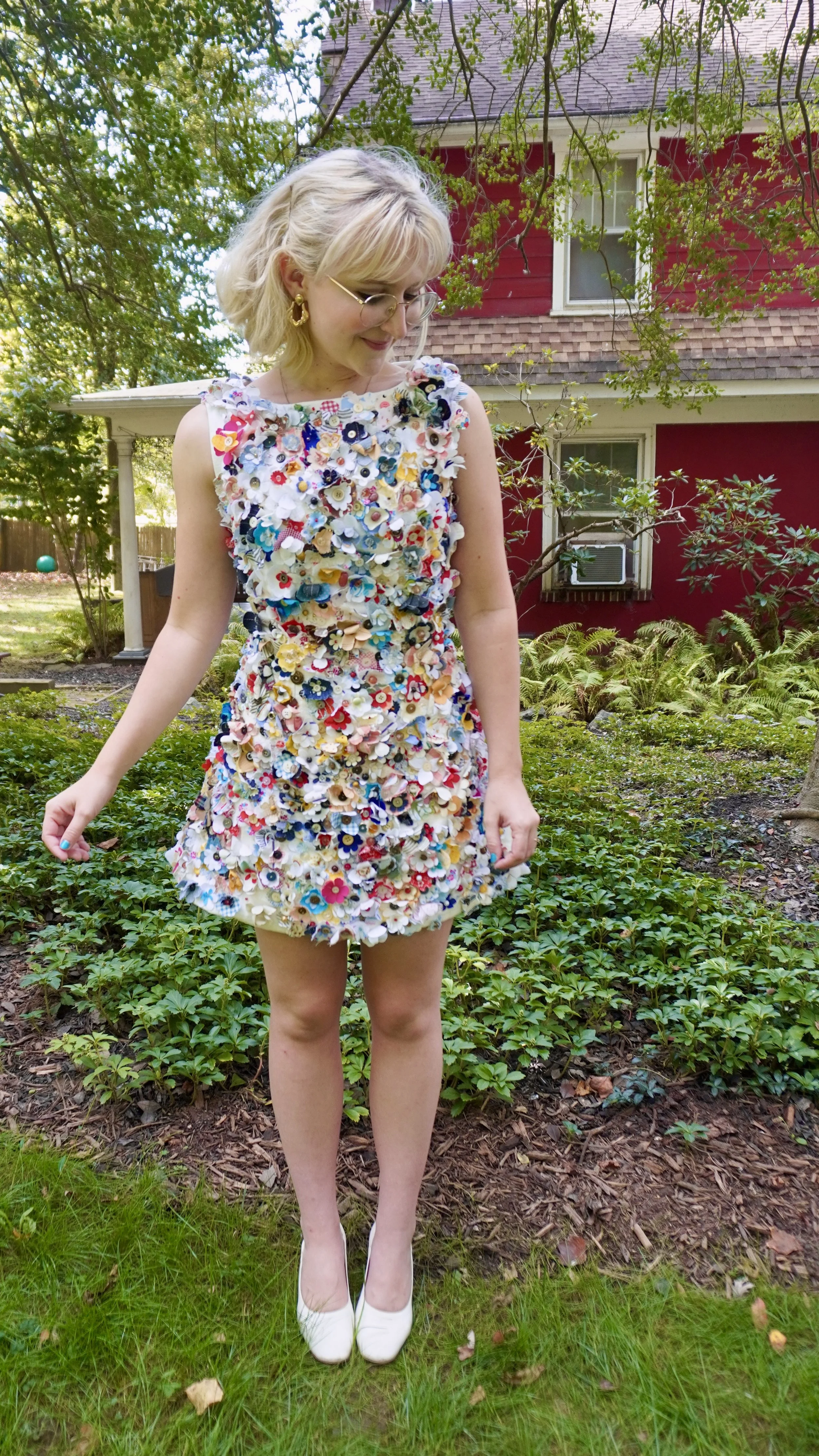 A woman standing outdoors in a garden, wearing a colorful dress made of various small fabric flowers, with a house in the background.