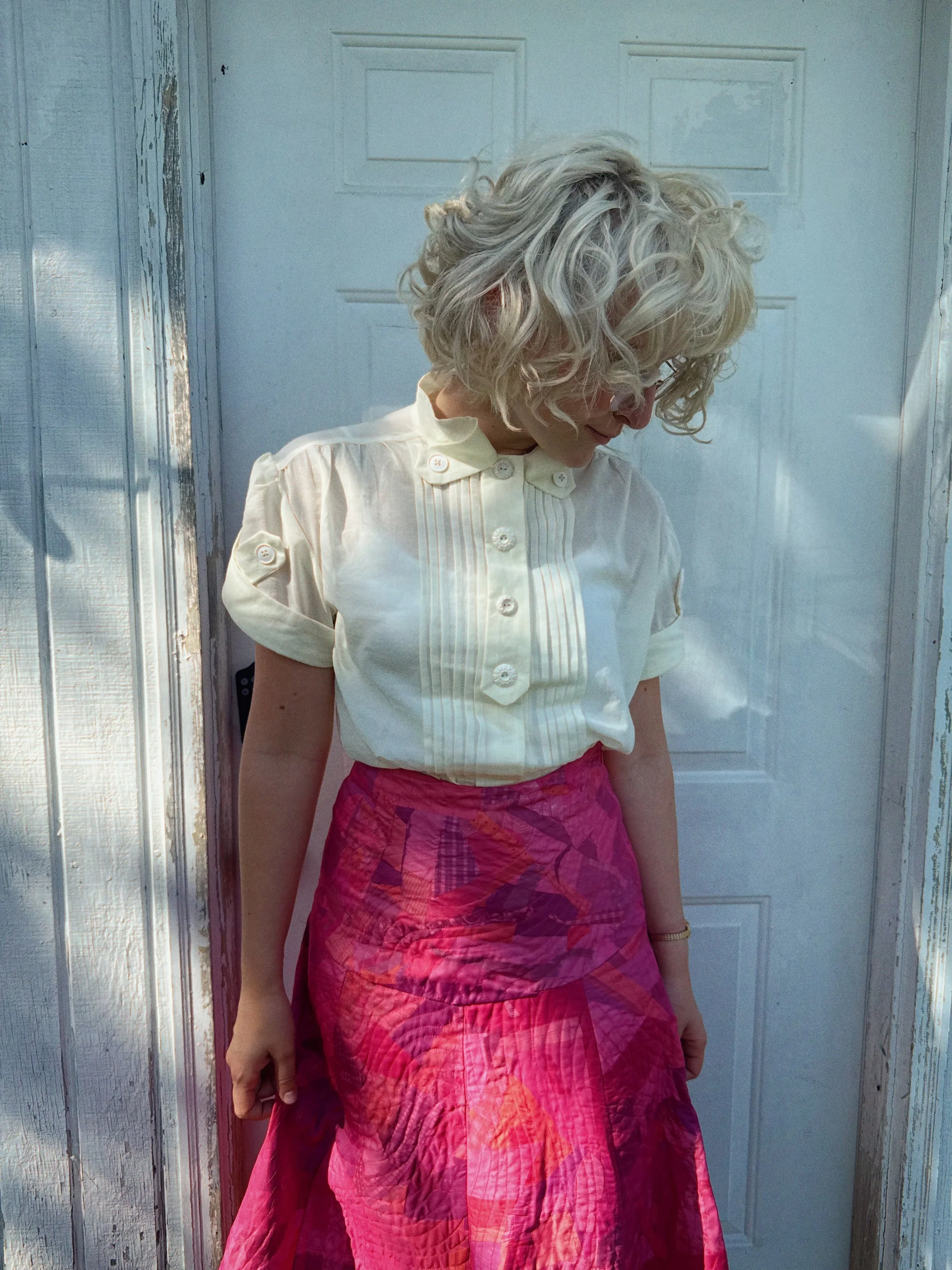 A woman with curly blonde hair wearing a white blouse and a pink skirt standing in front of a white door with weathered wooden trim.