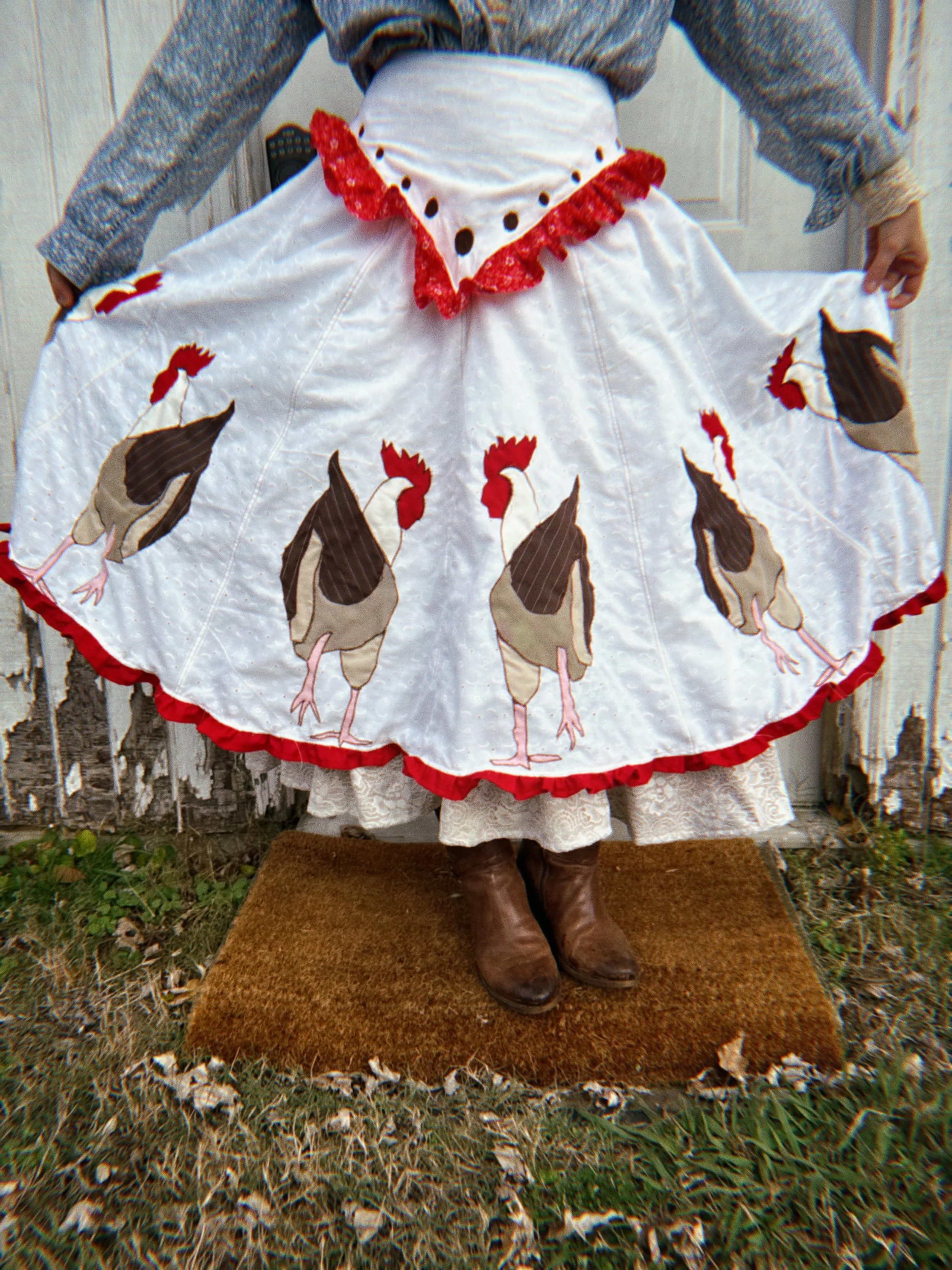 A person wearing boots and a white apron with rooster designs, exposing part of lace clothing underneath, standing on a brown carpet outside in front of a weathered fence.