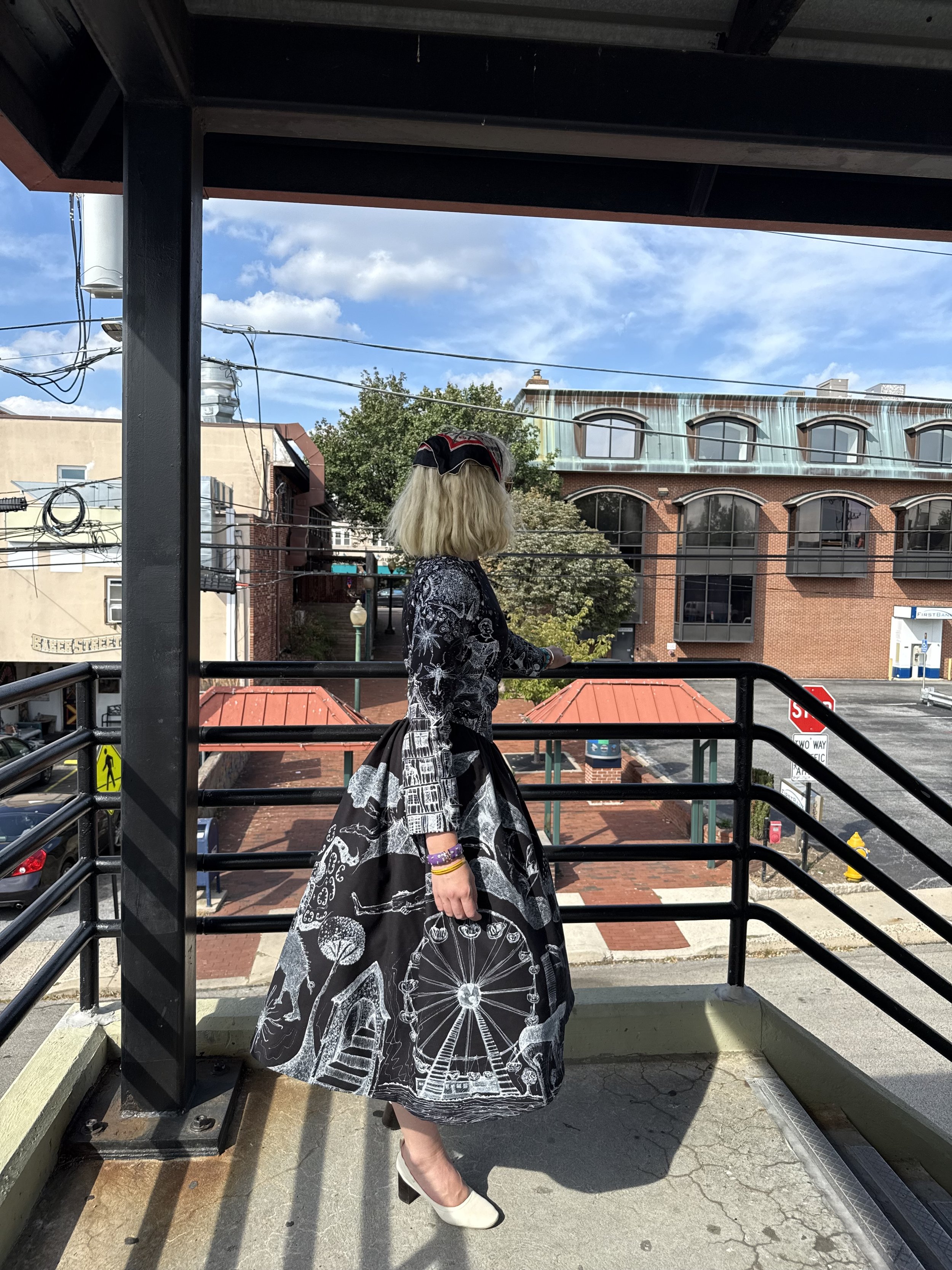 Woman with blonde hair wearing a black hat with red and white accents, black and white patterned dress, white shoes, standing on a balcony with black railing, overlooking a city street with brick buildings and a blue sky with clouds.