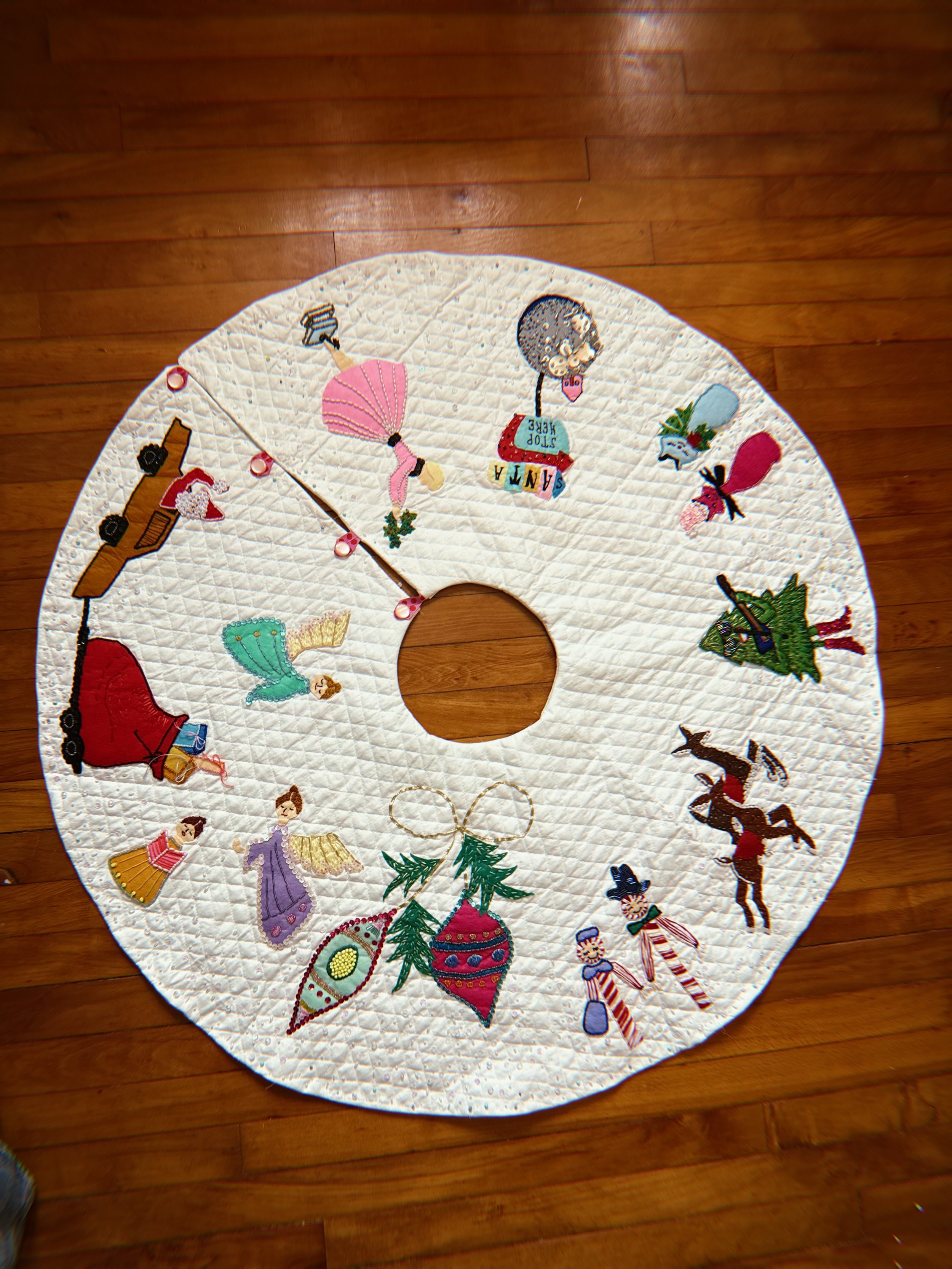 Circular quilt with Christmas-themed embroidery on a wooden floor. Embroidered figures include a Santa Claus, Christmas trees, angels, reindeer, elves, and holiday decorations.