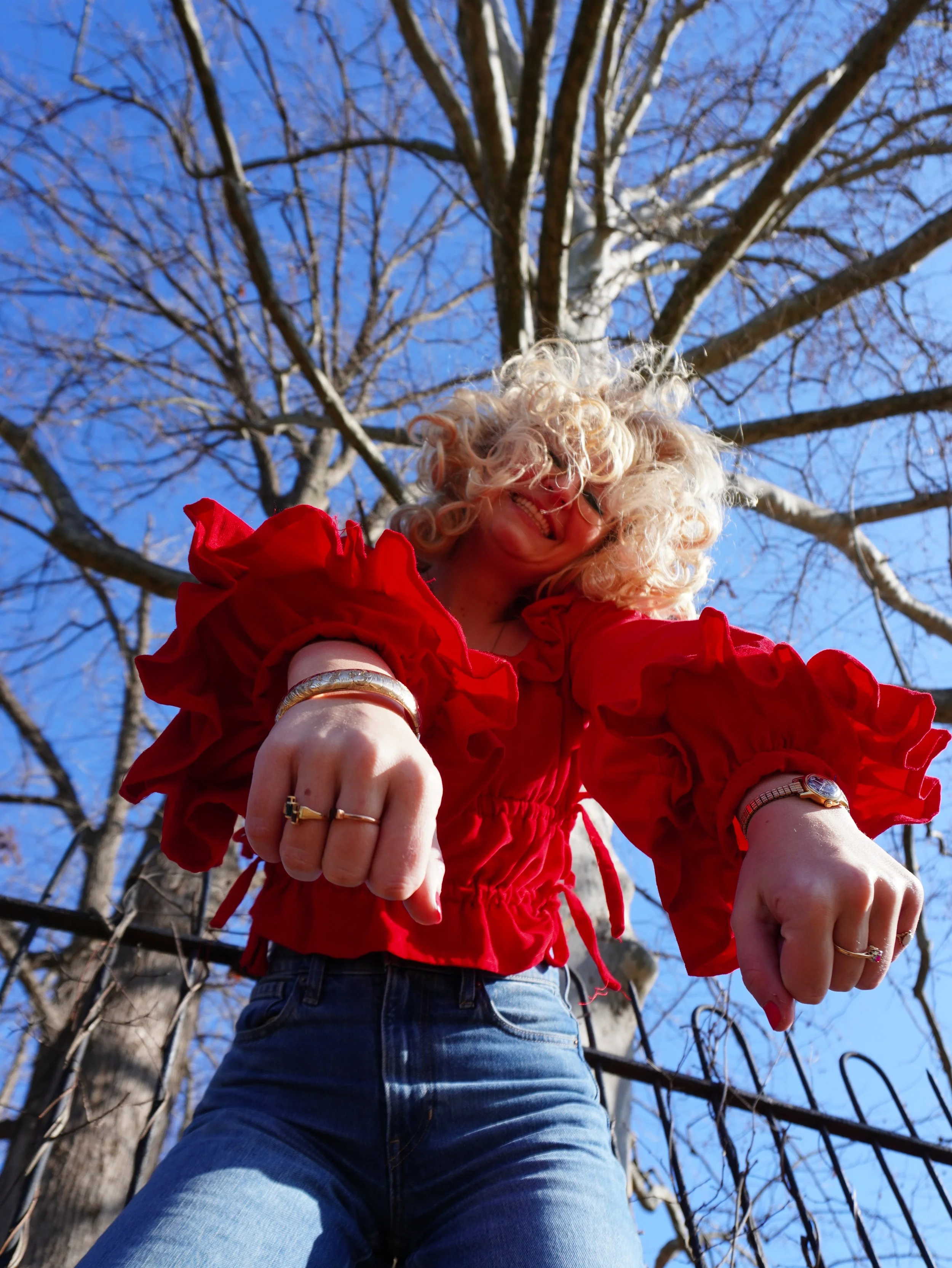 A woman with curly blonde hair and glasses wearing a red jacket and blue jeans is smiling and looking down at the camera, taken from a low angle outdoors with a blue sky and leafless tree in the background.