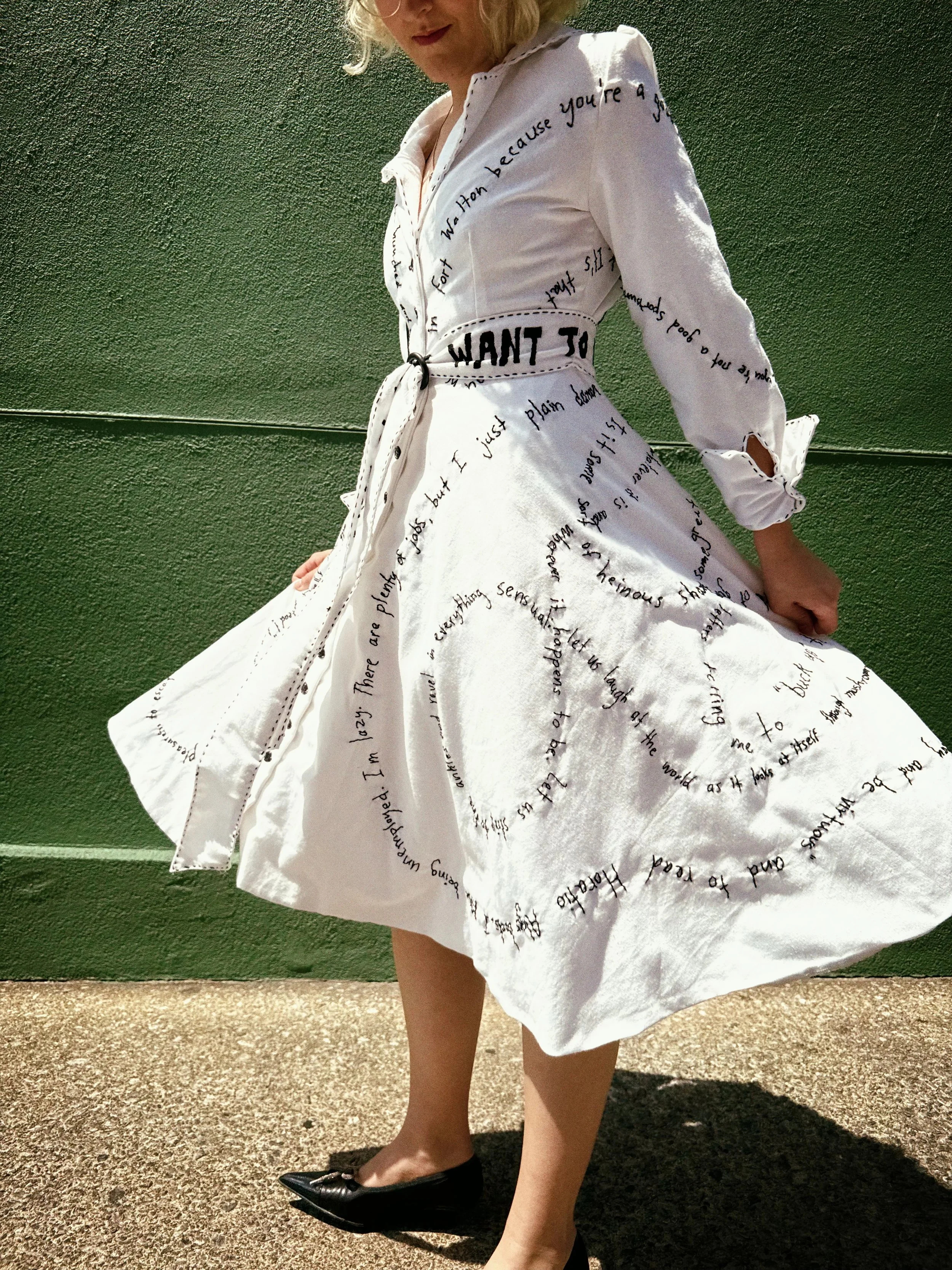 A woman wearing a white dress with black handwritten-style text and a black belt with the word 'WANT' printed on it. She is standing outdoors against a textured green wall, with her hand behind her back and wearing black high heels.