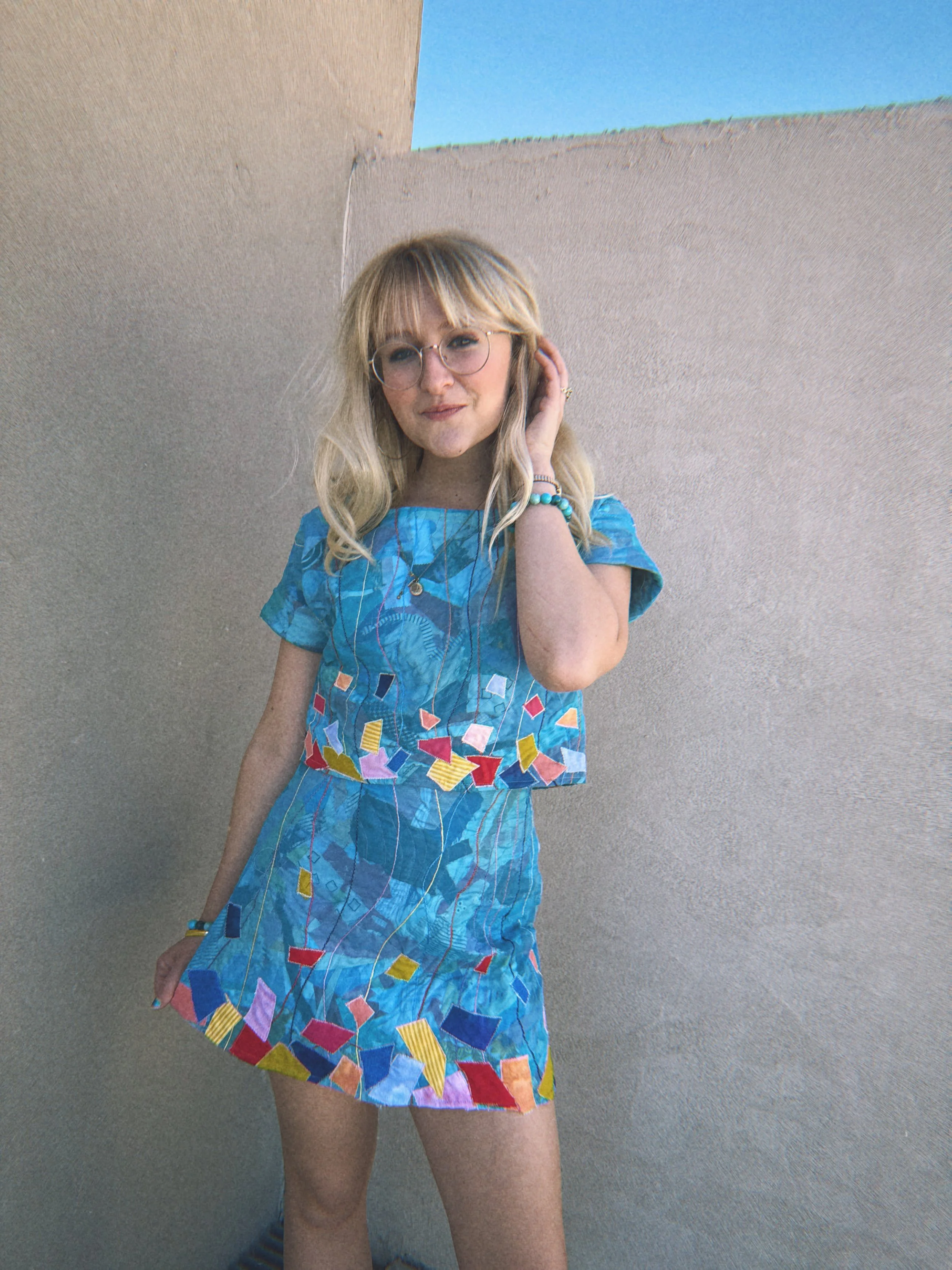 A young woman wearing glasses and a blue dress with colorful shapes, standing against a beige wall with an opening revealing blue sky.