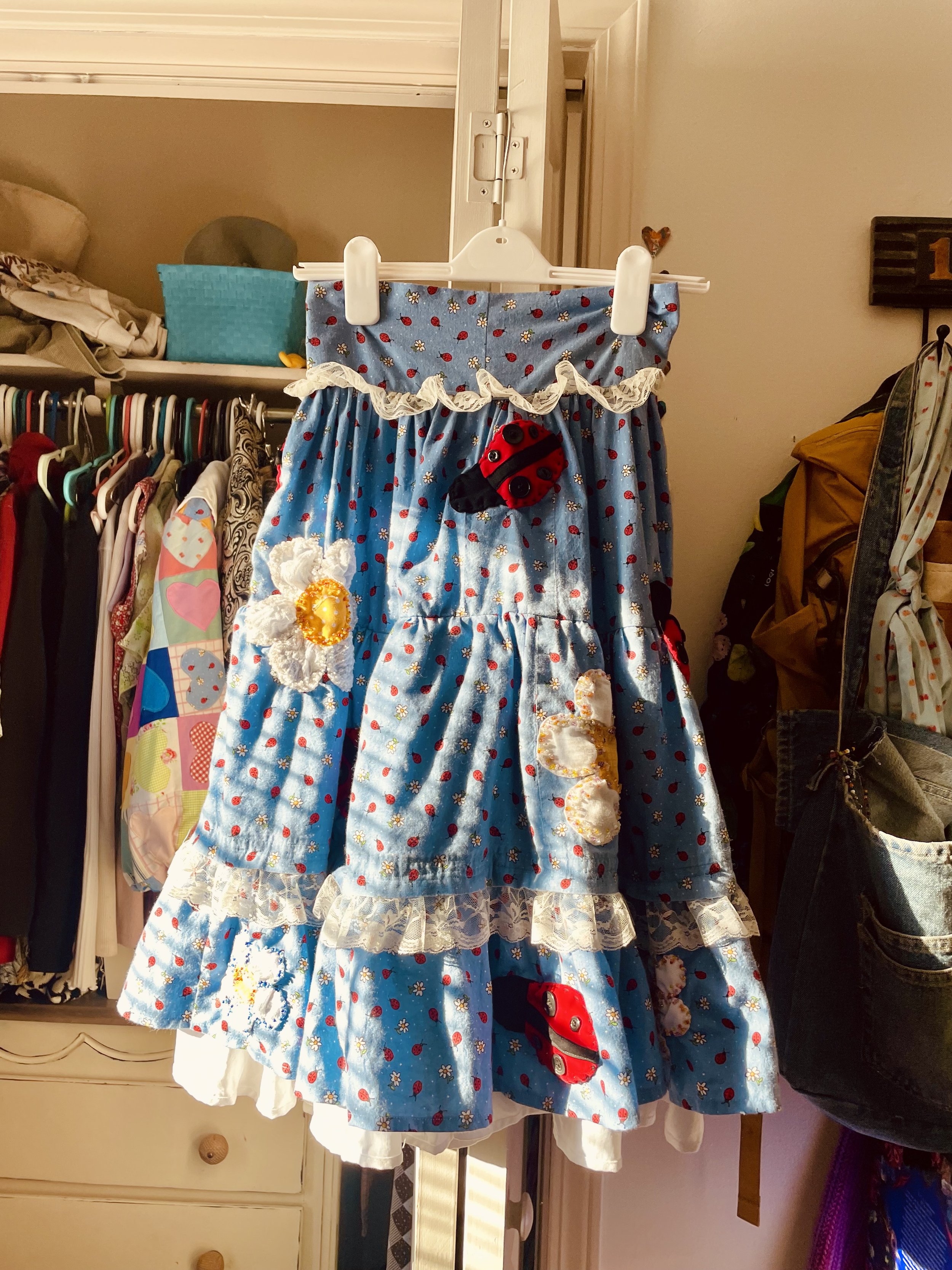 A blue dress with red and white floral pattern and lace trim, decorated with a plush ladybug, hanging on a white hanger in a closet.