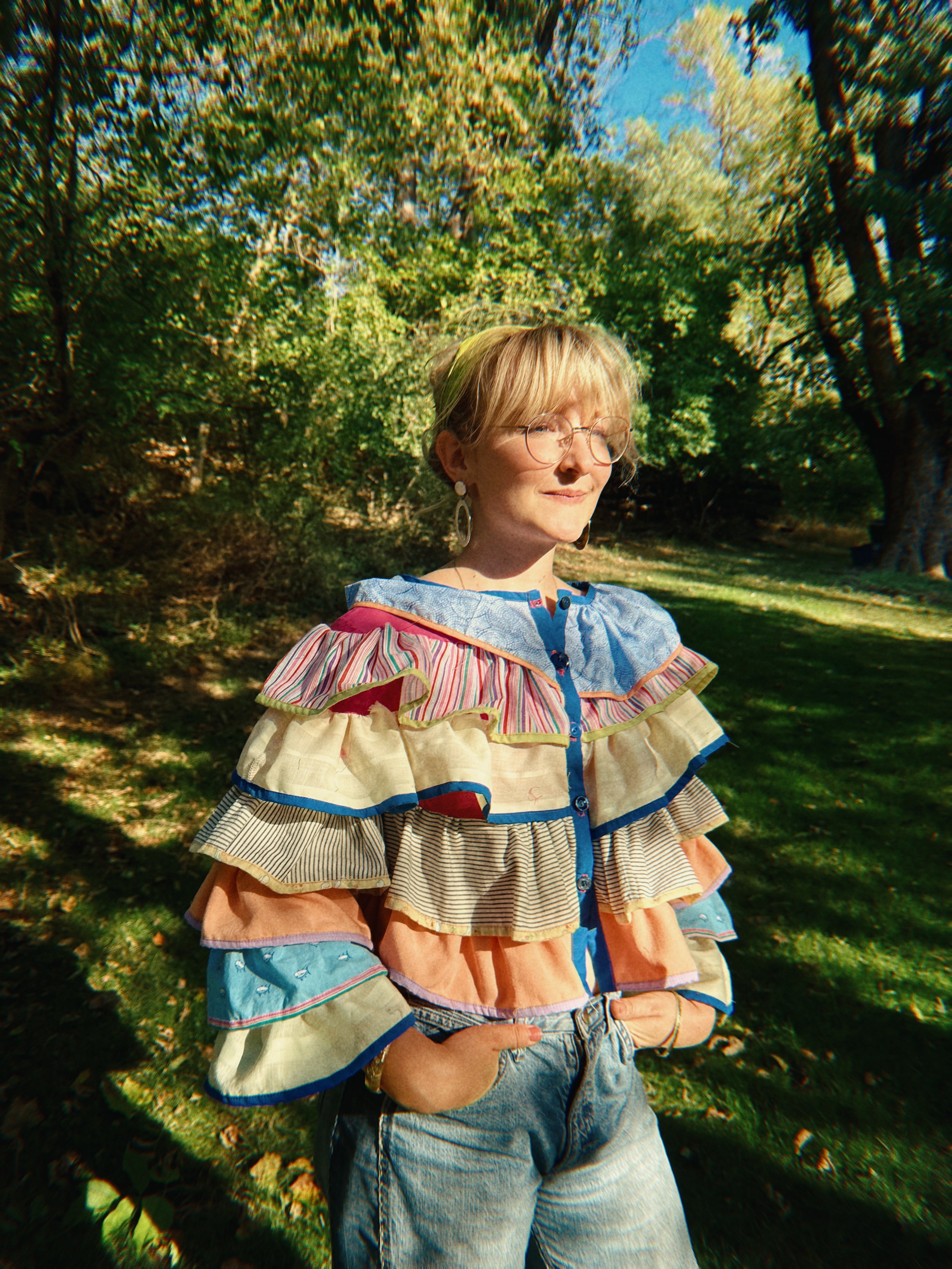 A woman with blonde hair and glasses standing outdoors in a sunny park with green trees in the background, wearing a colorful striped ruffled blouse and denim shorts.