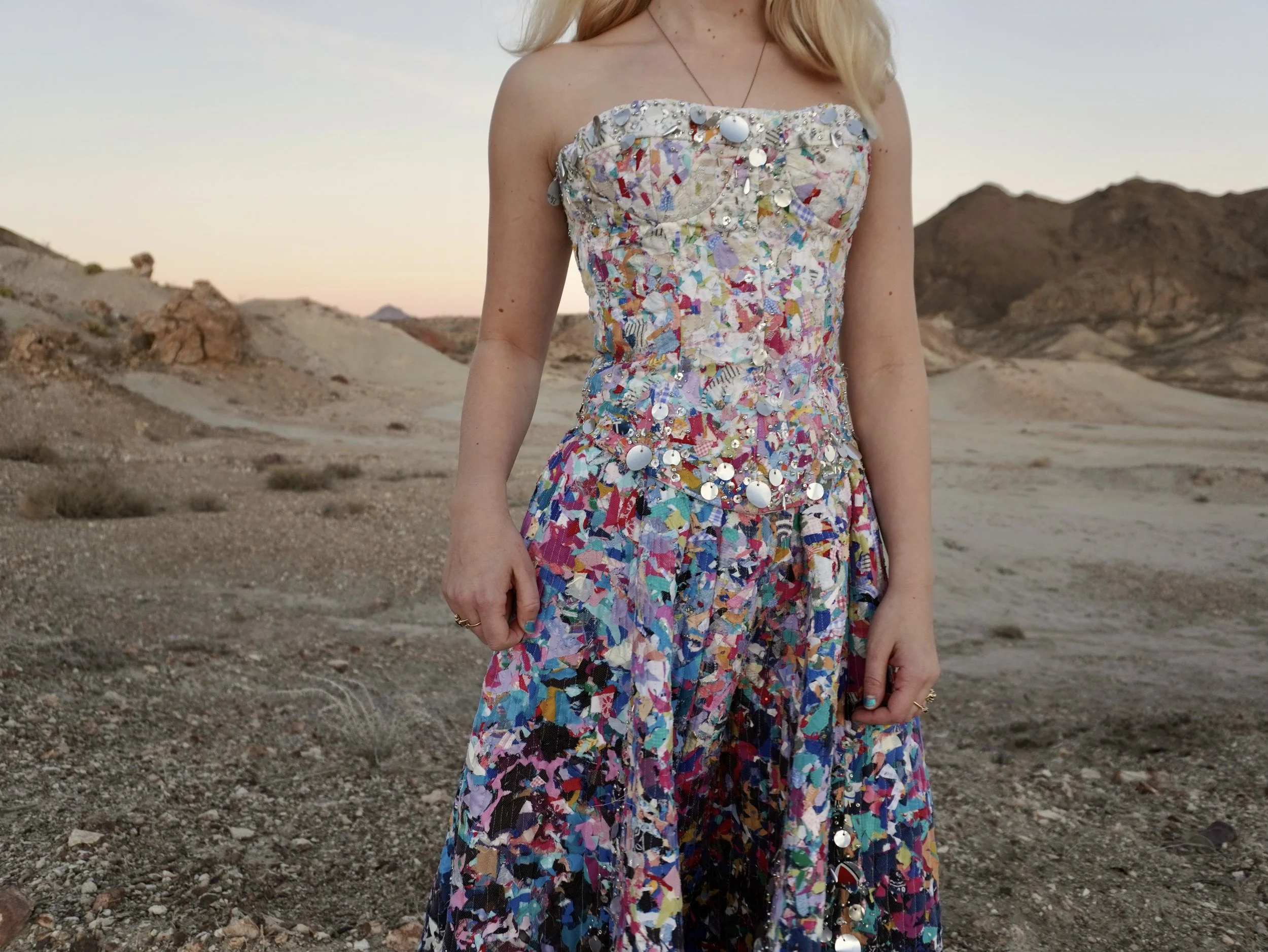 Close-up of a woman wearing a colorful, sequin-embellished dress, standing outdoors in a desert landscape during sunset.