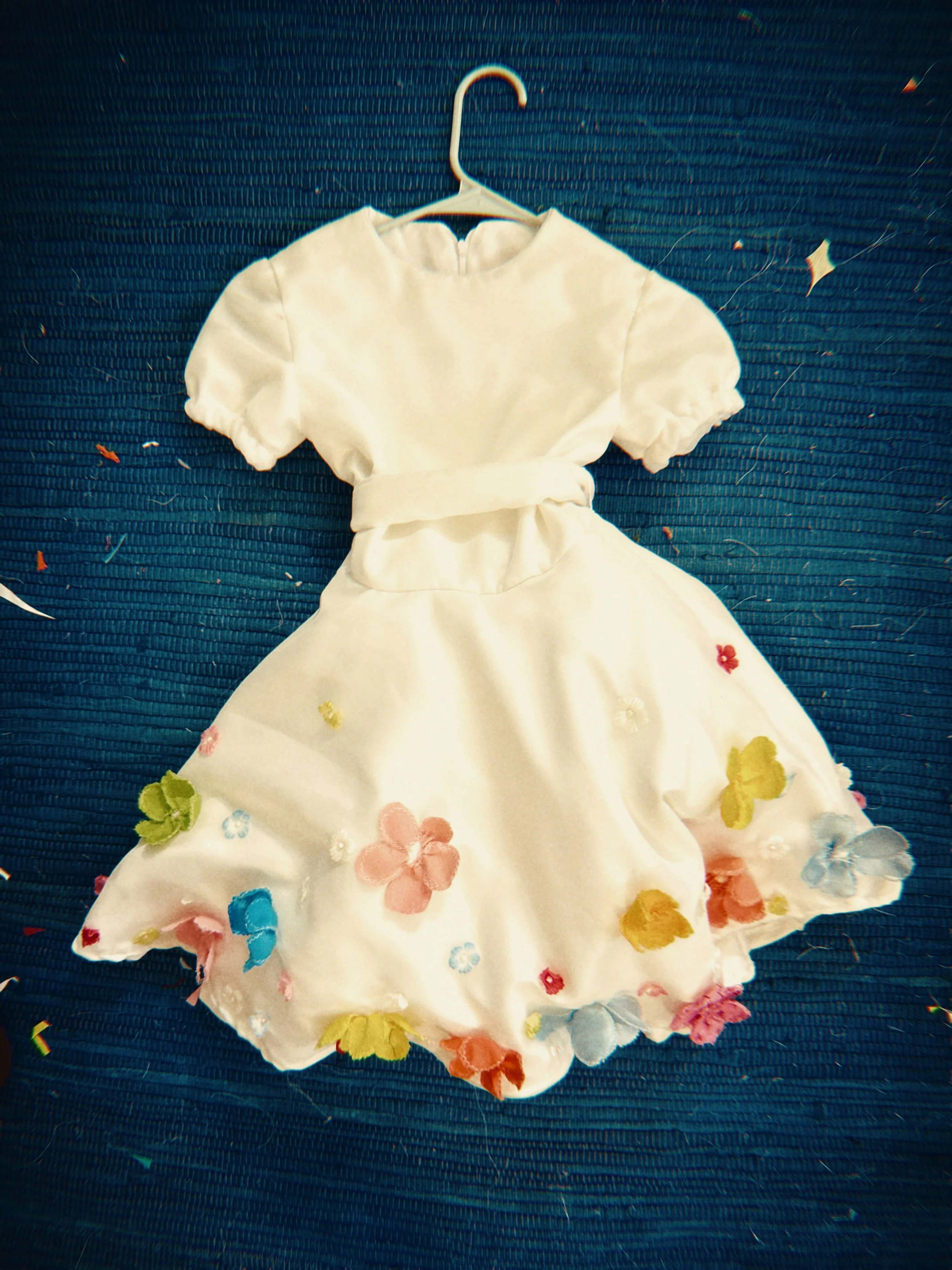 A white dress with colorful floral embroidery hanging on a white hanger against a dark blue textured background.