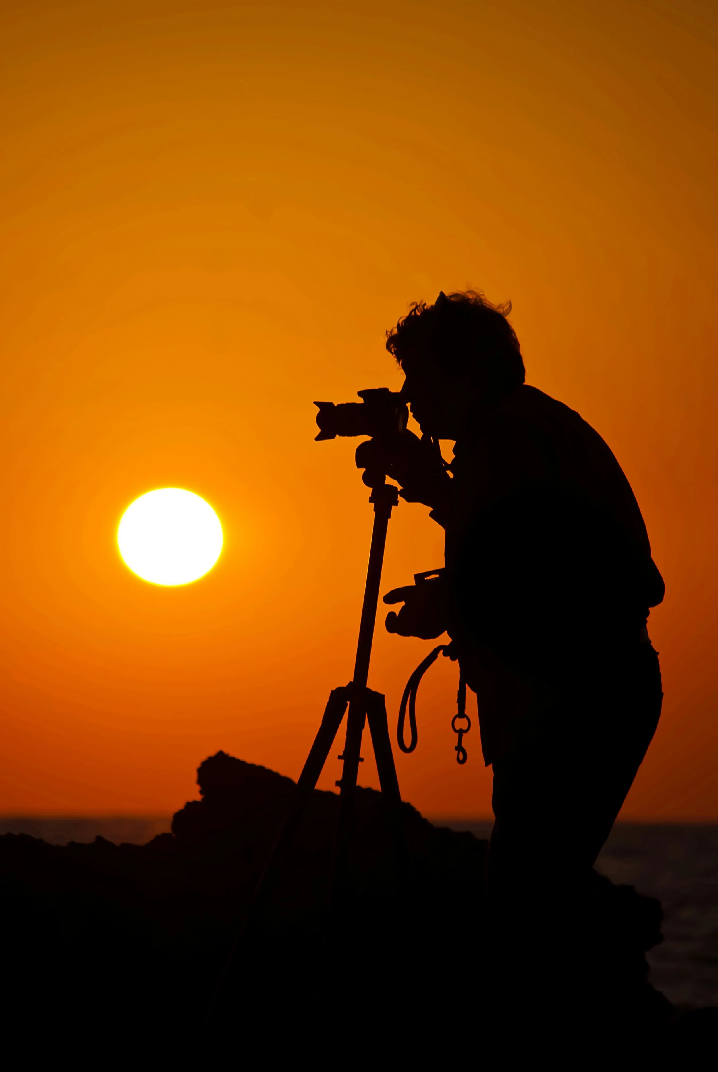 Silhouette of a person operating a camera on a tripod during sunset or sunrise over the ocean.