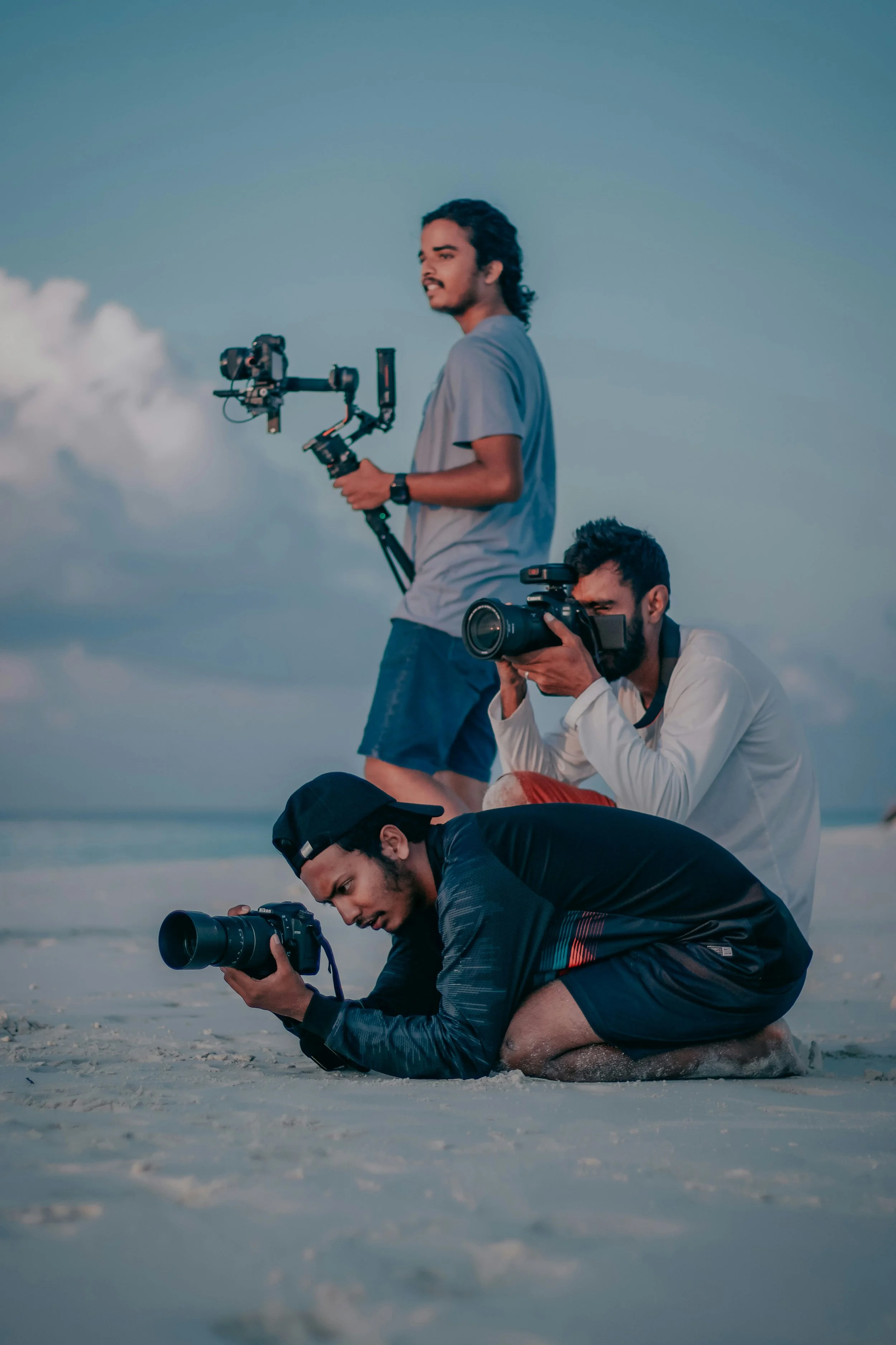 Three young men are on the beach, two are standing and one is kneeling, all holding cameras and filming or taking photos with cloudy sky in the background.