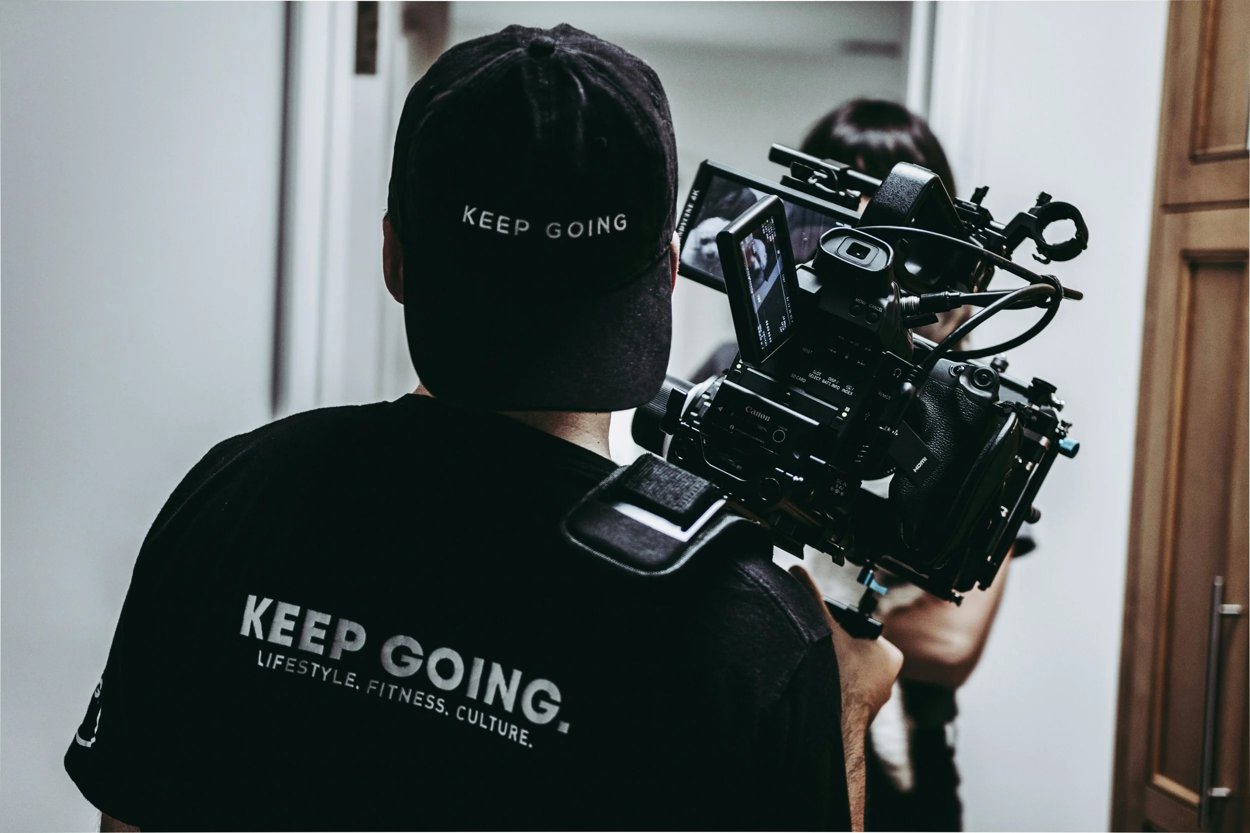 A person holding a professional camera with a mounted monitor, wearing a black cap and T-shirt that reads "Keep Going." standing in a room with white walls and wooden furniture, directing a scene with a woman.