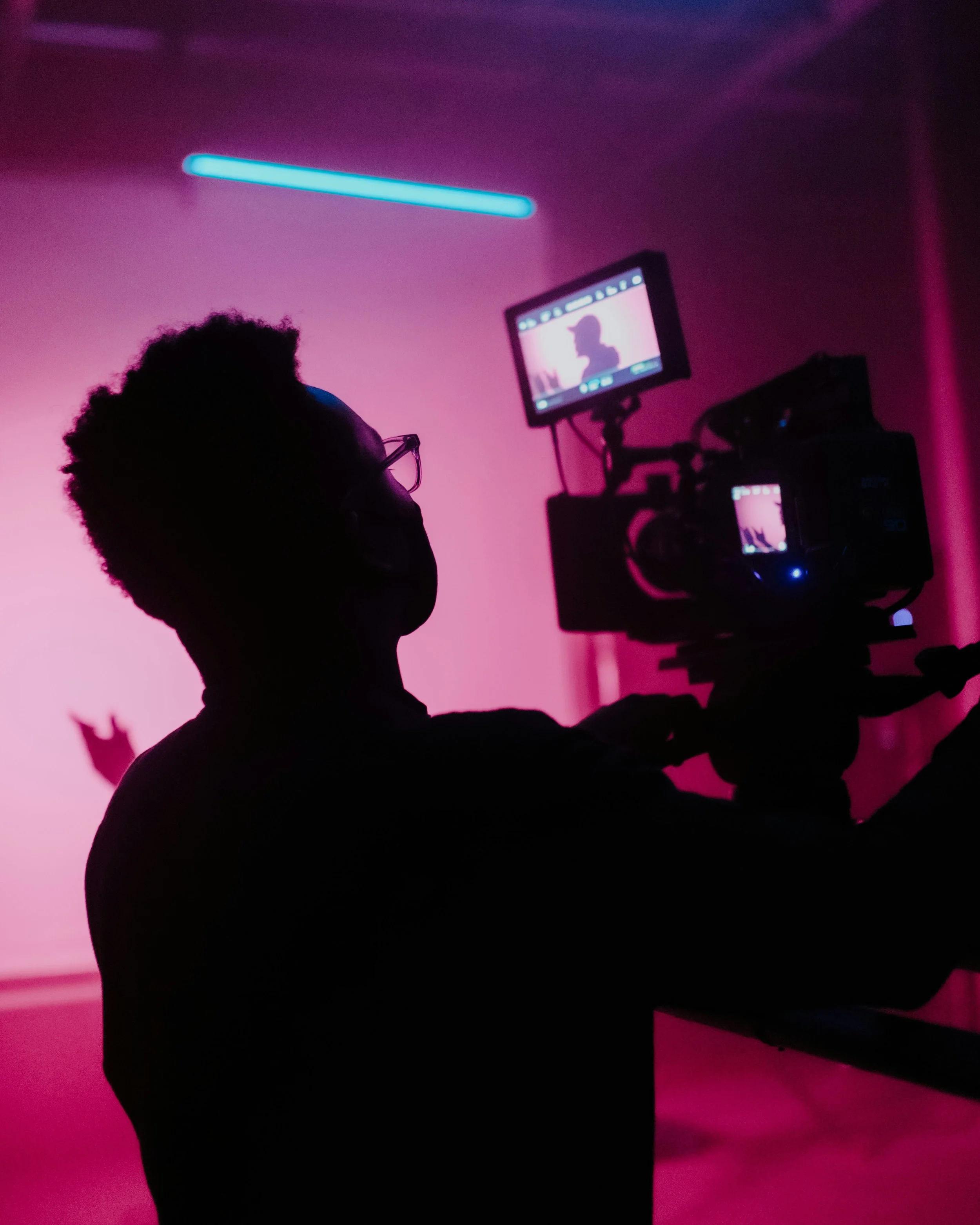 Silhouette of a person operating a camera in a pink and purple lit studio, with a monitor displaying a silhouetted scene.
