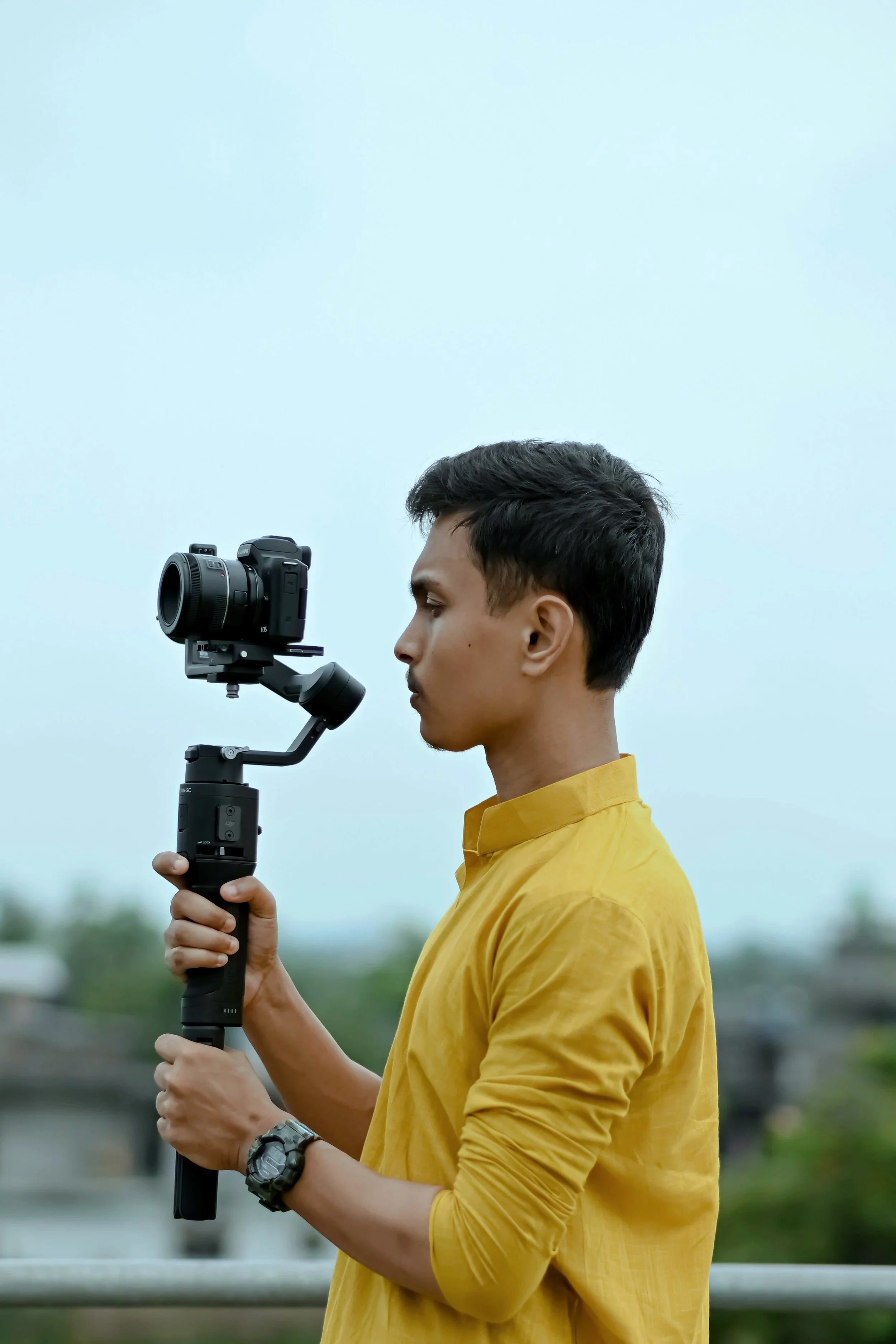 Young man in a yellow shirt holding a camera stabilizer with a camera attached, standing outdoors.