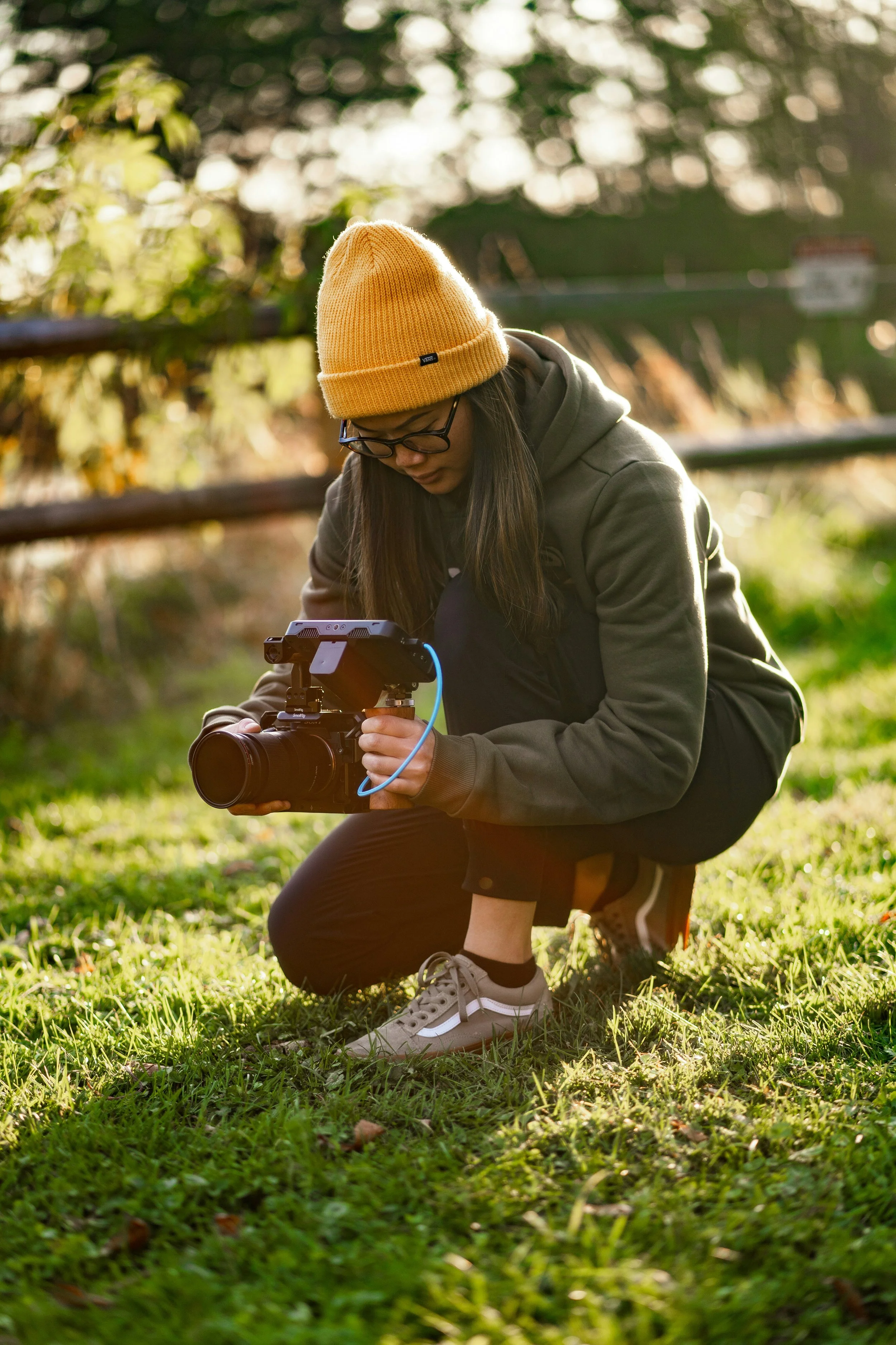 A person with long dark hair, glasses, a yellow beanie, a dark hoodie, black pants, and sneakers is crouching on grass while looking at a professional video camera mounted on a stabilizer in a park during late afternoon.
