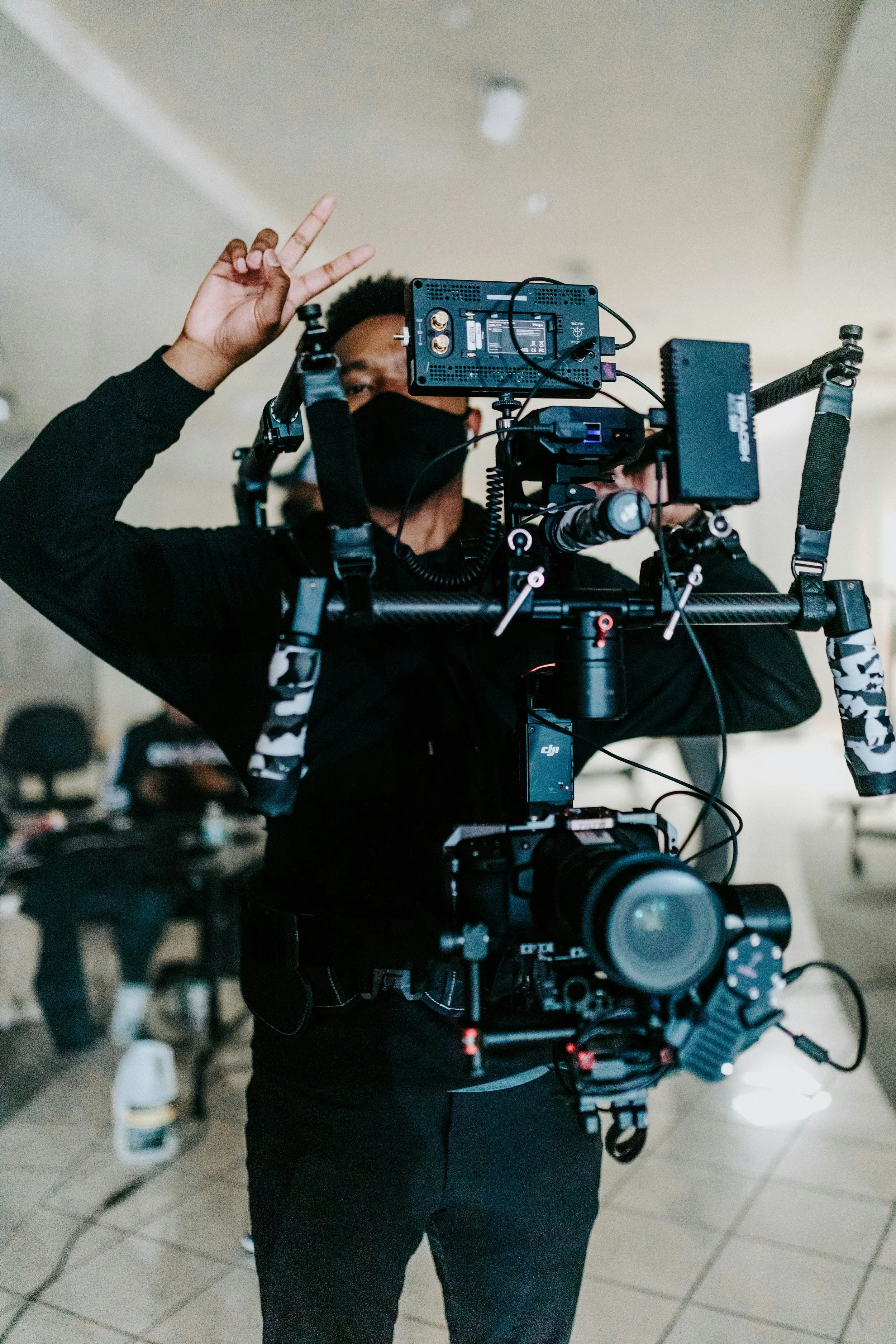 A person wearing a black mask and black clothing operating a professional camera mounted on a stabilizer rig, making a peace sign with their right hand.